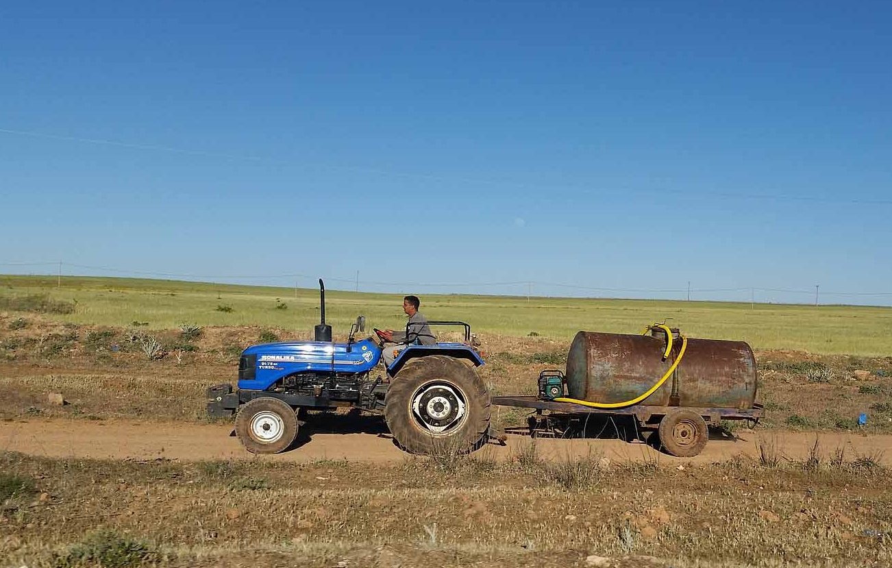 L'image montre un homme conduisant un tracteur bleu sur une route de terre. Il tire une remorque avec un grand réservoir métallique. À l'arrière-plan, on peut voir des champs verts sous un ciel dégagé. La scène évoque une ambiance rurale et agricole.