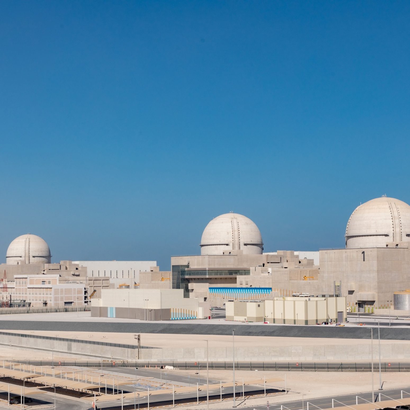 La imagen muestra una planta de energía nuclear con varias estructuras de reactor. Hay tres domos visibles en la parte superior de los reactores, que son características típicas de las centrales nucleares. La planta parece moderna y está rodeada de un entorno desértico, con un cielo claro y azul en el fondo. Hay edificios de soporte y áreas de estacionamiento en primer plano, lo que sugiere que es una instalación grande y compleja.
