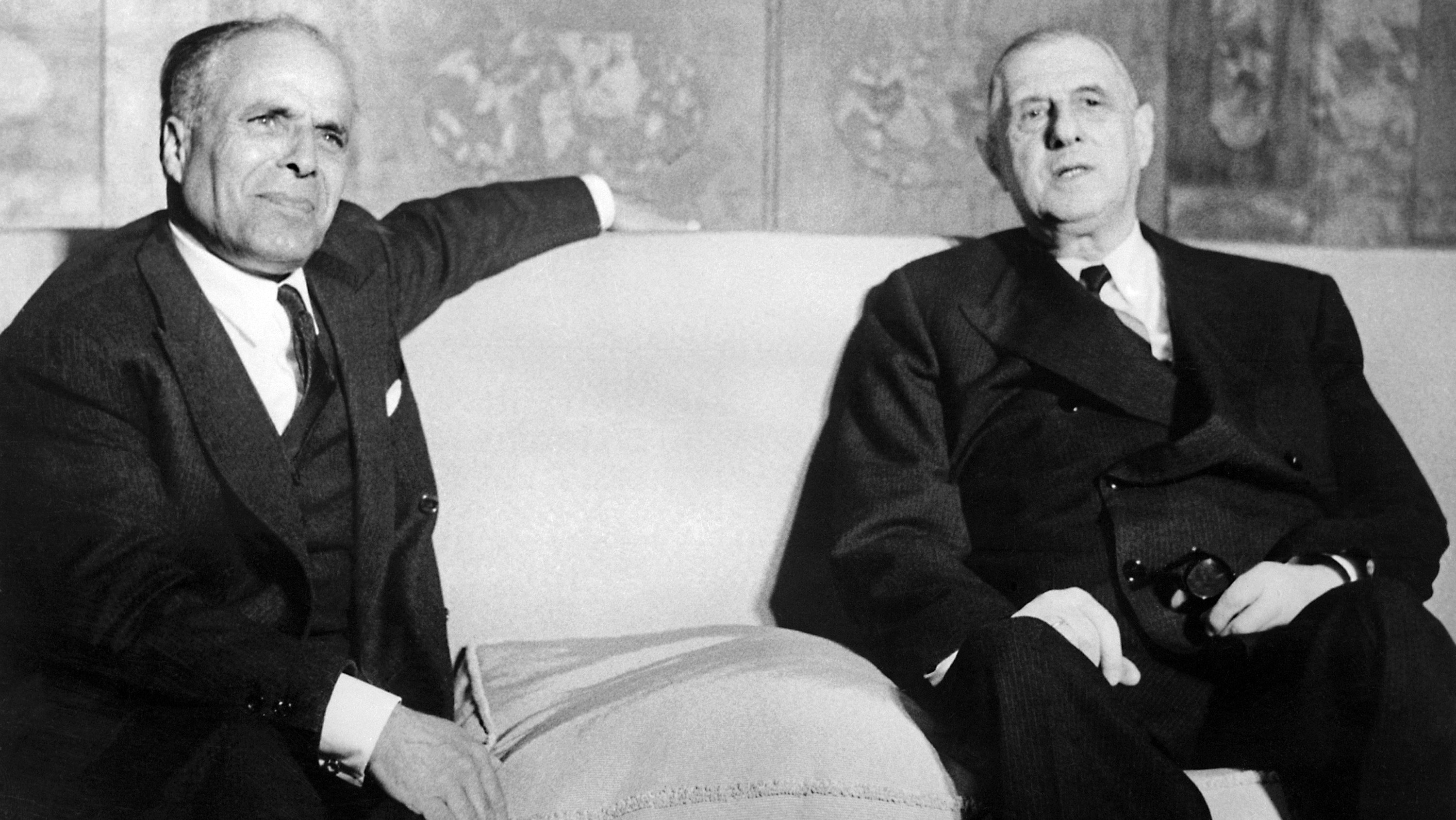 L'image montre deux hommes assis sur un canapé dans un environnement formel. Ils ont l'air sérieux et portent des costumes sombres. L'un des hommes semble légèrement se pencher en arrière, tandis que l'autre regarde devant lui. L'arrière-plan est décoré, mais les détails ne sont pas clairement visibles. L'atmosphère générale apparaît académique et solennelle.