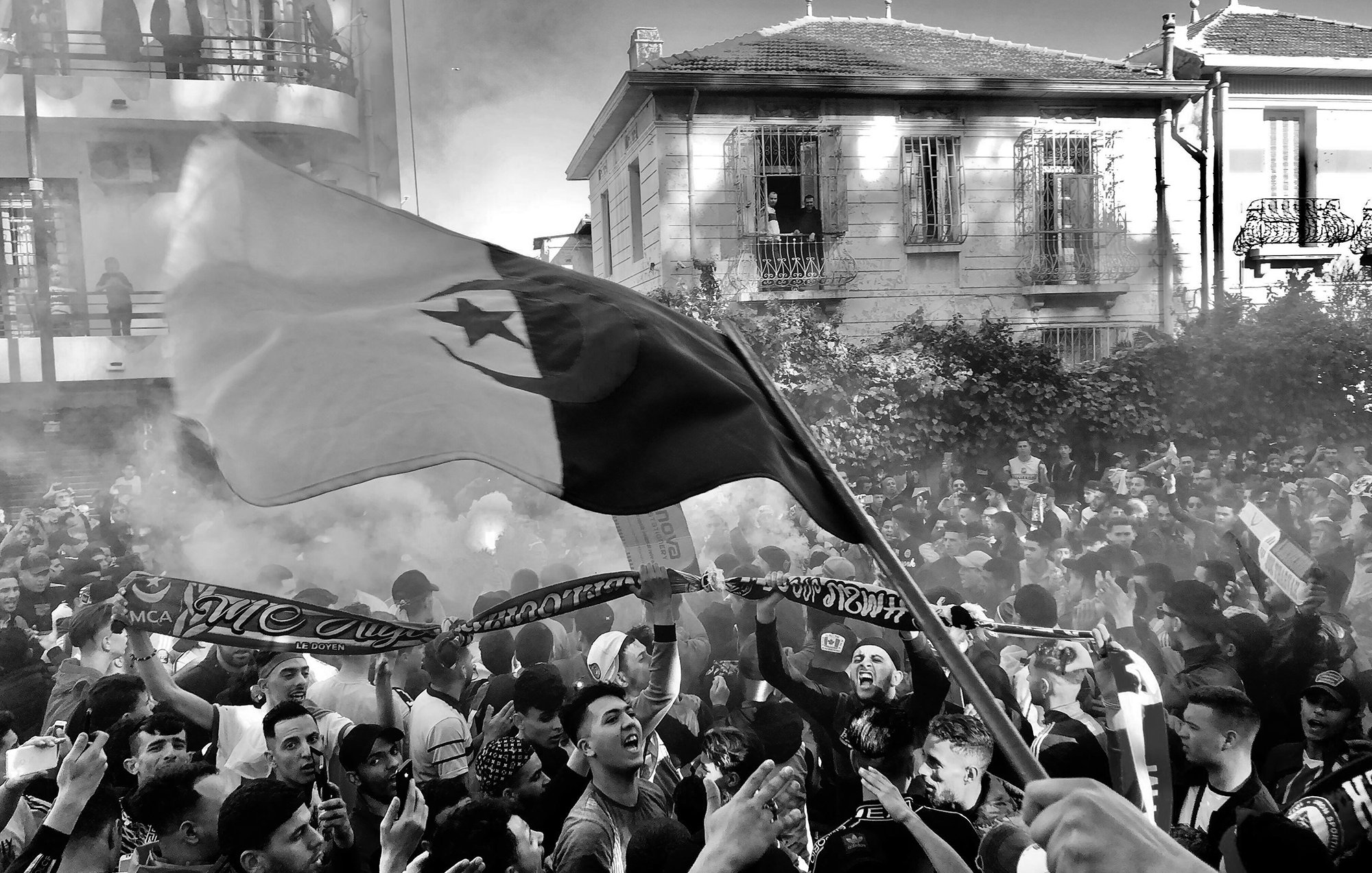 L'image montre une scène vibrante et animée, probablement lors d'un événement festif ou d'une manifestation. On peut voir une grande foule de personnes rassemblées, certaines brandissant des drapeaux et des écharpes. Les visages expriment la joie et l'enthousiasme. Une atmosphère de célébration est accentuée par des fumigènes qui créent un léger brouillard. En arrière-plan, on distingue des bâtiments, ce qui suggère que cette scène se déroule en milieu urbain.