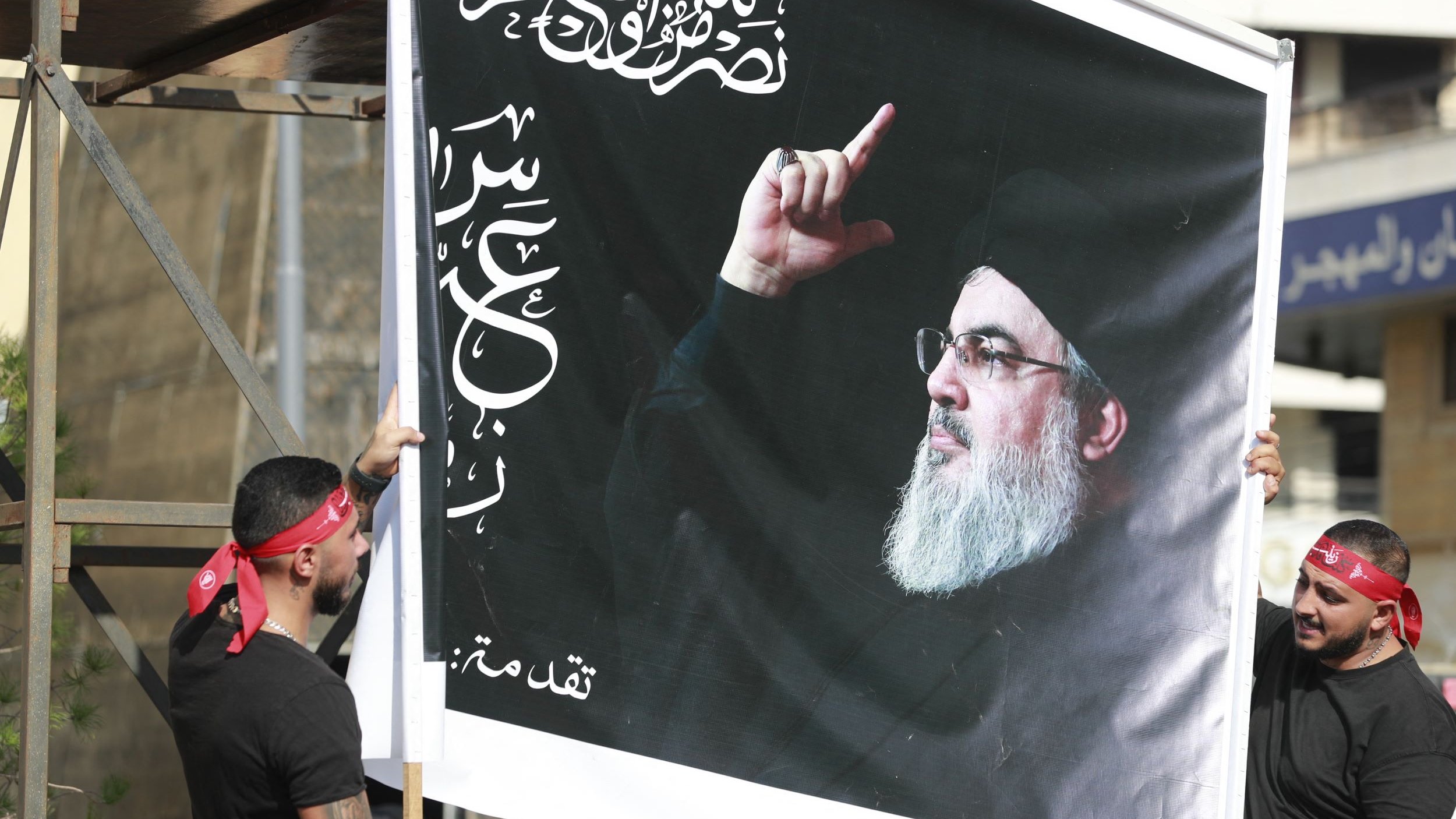 L'image montre deux personnes tenant une grande affiche. Cette affiche représente un homme avec une barbe, portant un vêtement sombre, et levant un doigt. En arrière-plan, on aperçoit des bâtiments. Les deux personnes portent des bandanas rouges et des t-shirts noirs. L'affiche comporte également des inscriptions arabes.