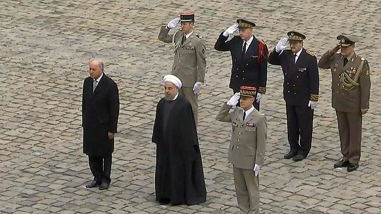 L'image montre plusieurs personnes en uniforme militaire saluant. Au centre, deux hommes en costume : l'un à gauche semble être un dirigeant, tandis que l'autre, portant une robe noire et un turban, est probablement un haut responsable religieux ou politique. Les autres personnes en arrière-plan sont des officiers en uniforme, également en train de rendre les honneurs. Ils se trouvent sur une surface en pavés, suggérant une cérémonie officielle ou un événement diplomatique.