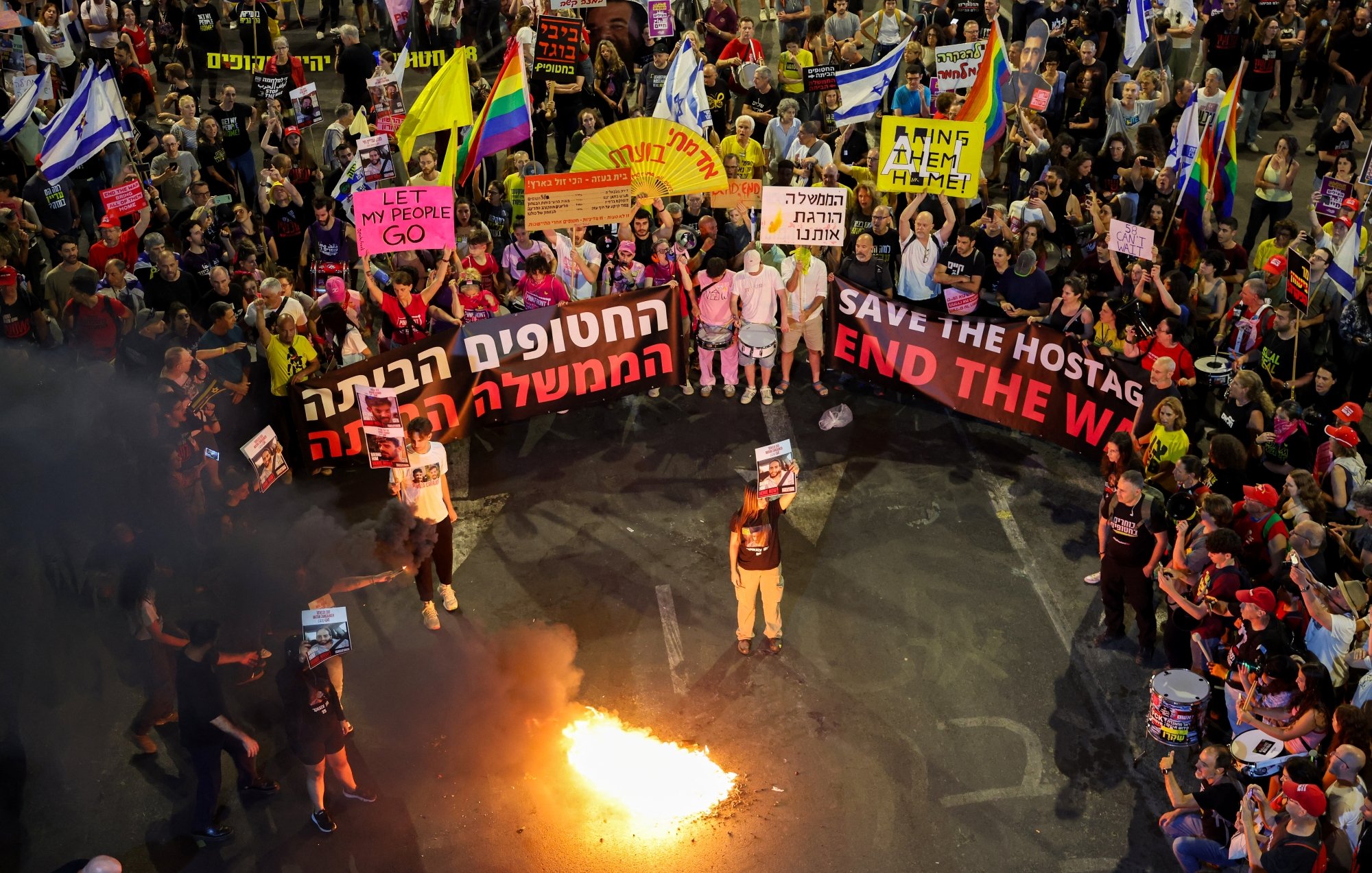 A large protest with colorful banners, people holding signs, and a fire in the center.