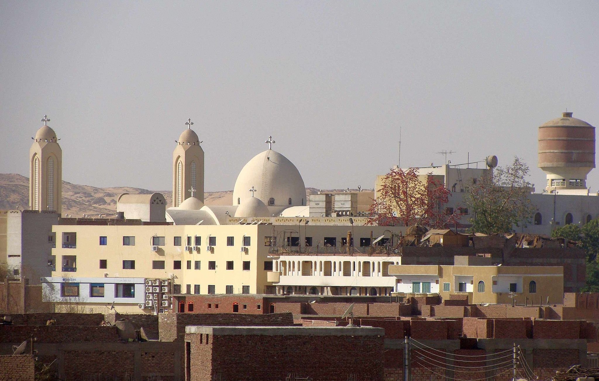 الصورة تظهر مجموعة من المباني في منطقة حضرية، حيث يتواجد مباني ذات طراز معماري مميز، بما في ذلك الكنائس التي تتضمن قبابًا وجرسًا. الخلفية تحتوي على تلال وصحراء، مما يوحي بأن المنطقة قد تكون قريبة من الطبيعة. يوجد أيضًا مجموعة من المباني السكنية المنخفضة، تظهر على اليمين، مما يعطي انطباعًا عن الحياة اليومية في تلك المنطقة.