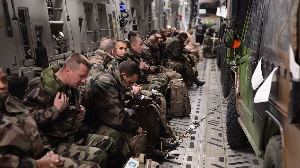 L'image montre un groupe de soldats assis à l'intérieur d'un véhicule militaire ou d'un avion de transport. Ils portent des uniformes camouflage et sont équipés de sacs à dos. Certains semblent attentifs et concentrés, tandis que d'autres sont plus détendus. Le cadre est sombre et fonctionnel, typique des espaces militaires, avec des sièges alignés sur les côtés et un véhicule militaire garé à l'intérieur.