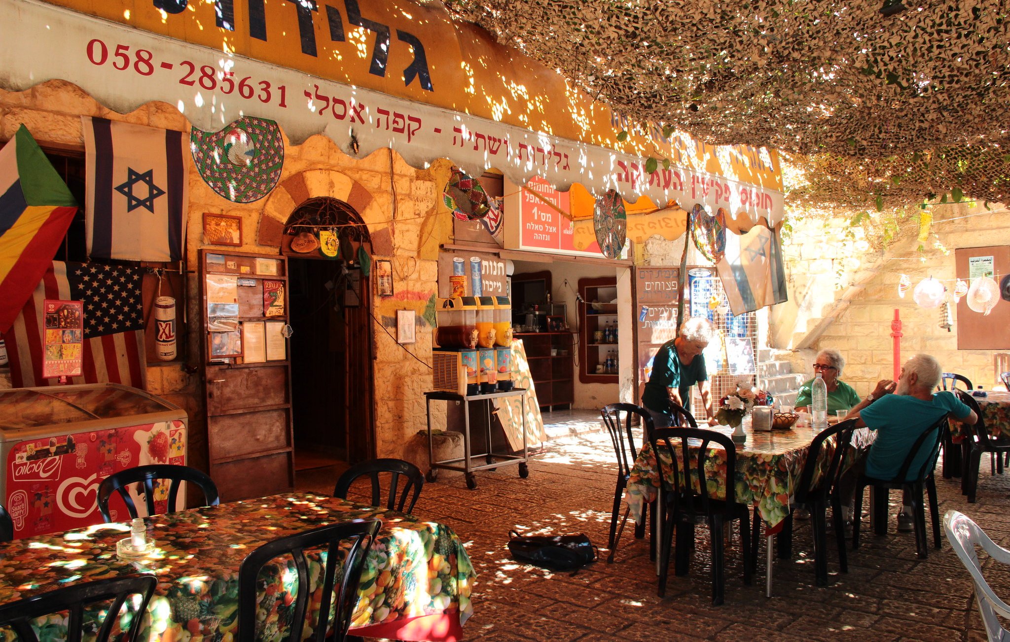 La imagen muestra un ambiente acogedor y colorido de un café o restaurante al aire libre. Hay mesas y sillas negras distribuidas sobre un suelo pavimentado, y la decoración incluye manteles con estampados florales. En el fondo, se pueden ver varias banderas, incluyendo la de Israel, colgadas en las paredes de piedra. El espacio está cubierto por una especie de toldo que filtra la luz, creando un ambiente agradable. Algunas personas están sentadas en las mesas, disfrutando del lugar.