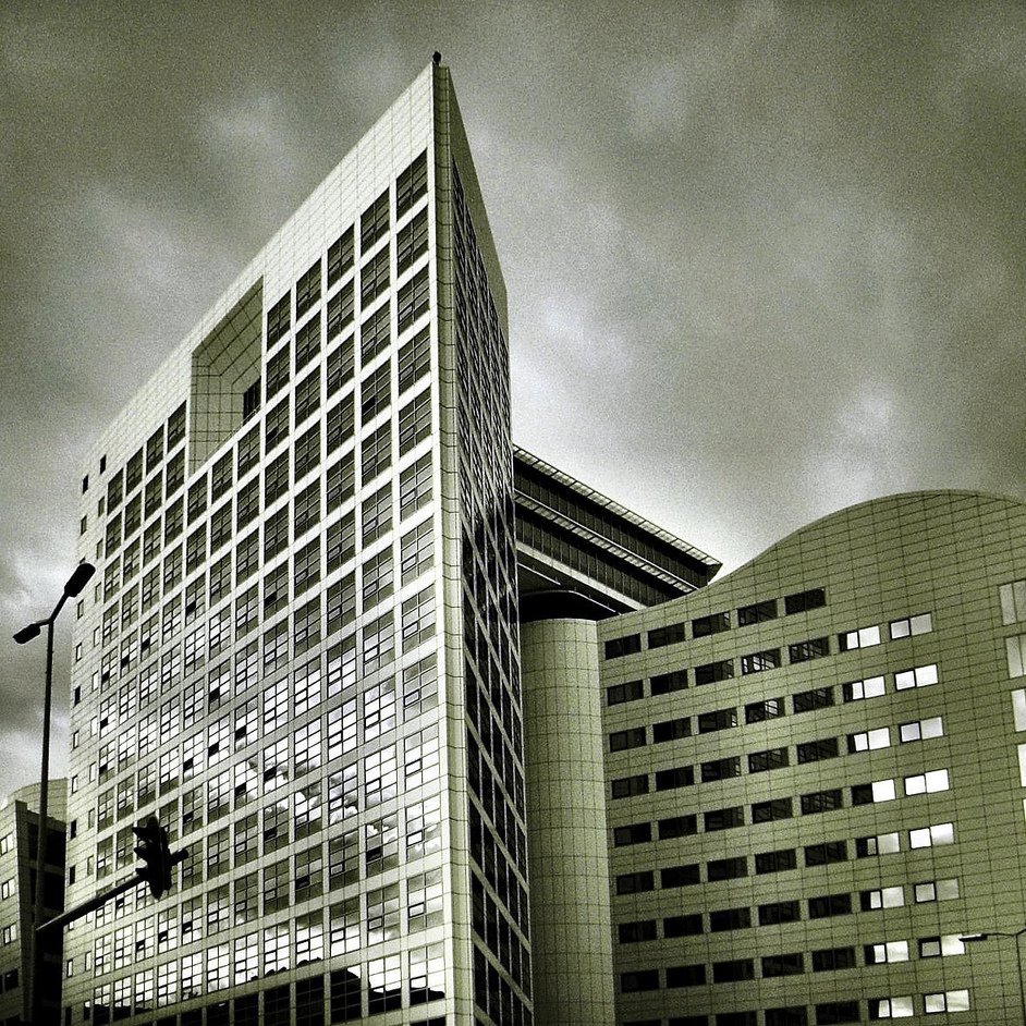 L'image montre un bâtiment moderne avec une architecture contemporaine. La structure est composée de plusieurs angles et surfaces réfléchissantes, donnant une impression de sophistication. Les vitres sont grandes, et le design présente des lignes épurées. Le ciel en arrière-plan est nuageux, ce qui peut créer une ambiance dramatique. Les tons de l'image sont en noir et blanc, accentuant les contrastes entre la lumière et l'ombre sur la façade du bâtiment.