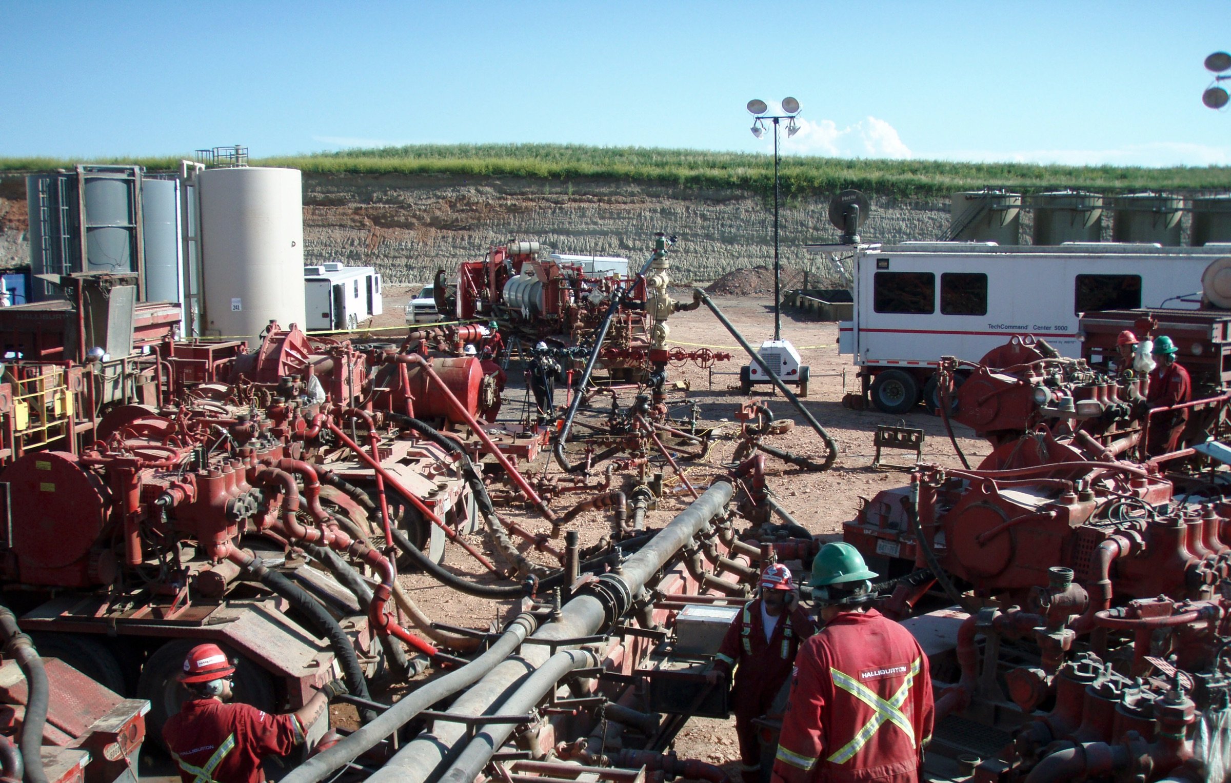L'image montre un site d'exploitation, probablement lié à l'industrie pétrolière ou gazière. On y voit plusieurs équipements imposants, notamment des pompes et des tuyaux, ainsi que des travailleurs en tenue de sécurité, portant des casques et des vêtements rouges. Le paysage en arrière-plan est rural, avec une colline et des installations industrielles. Le ciel est dégagé, laissant suggérer une journée ensoleillée. L'ensemble de la scène évoque une activité intense et technique, généralement associée à des opérations d'extraction ou de forage.