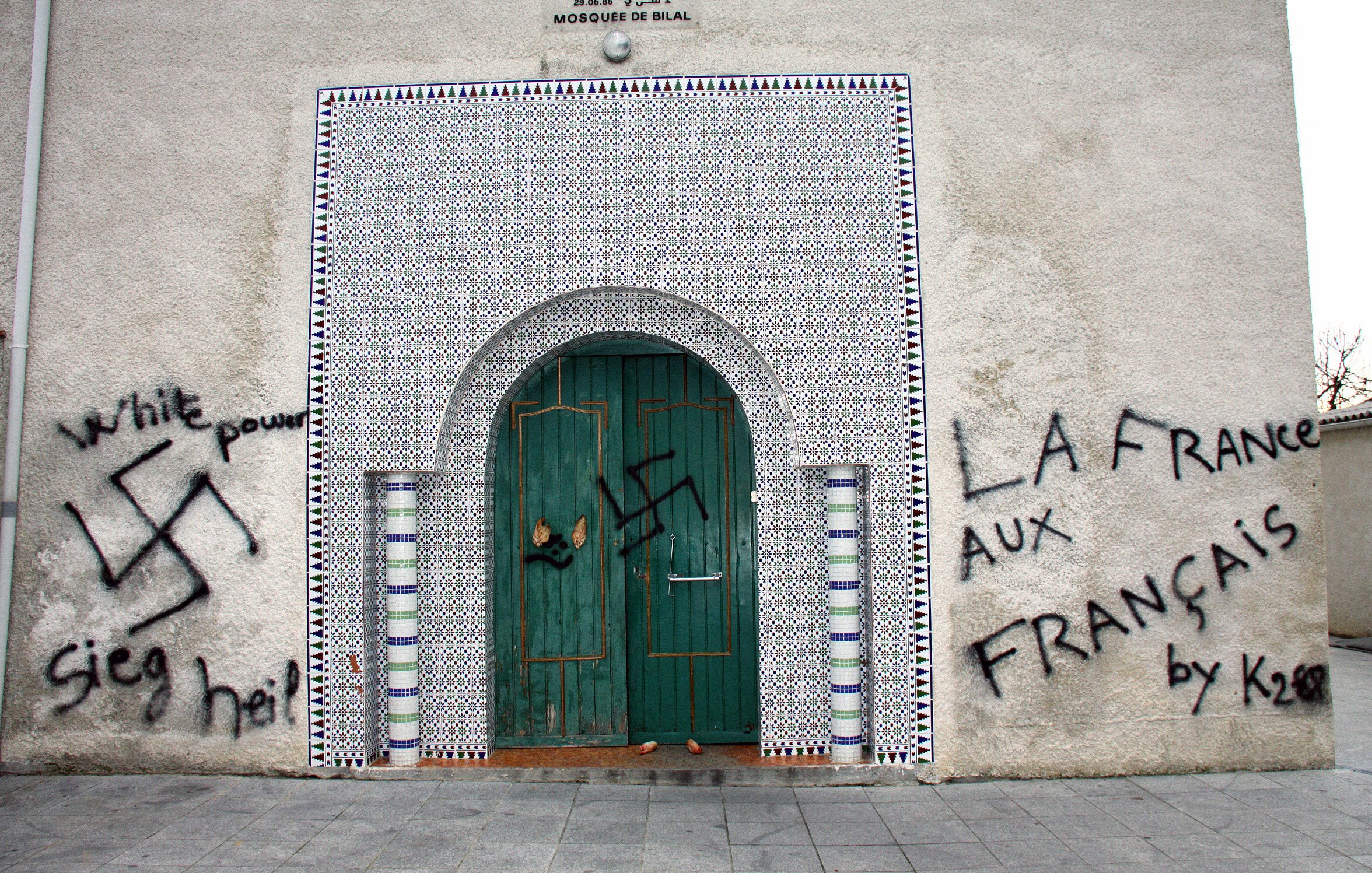 این تصویر نمایی از دیواری را نشان می‌دهد که در آن نمادهای نژادپرستی و خشونت، از جمله علامت هیکل و نوشته‌هایی به زبان‌های مختلف، Spray شده است. در بالای درب ورودی، طراحی‌های اسلامی به چشم می‌خورد که نشان‌دهنده یک مسجد است. دیوار به طور کلی نشان‌دهنده تضاد بین هنر و پیام‌های نفرت است.