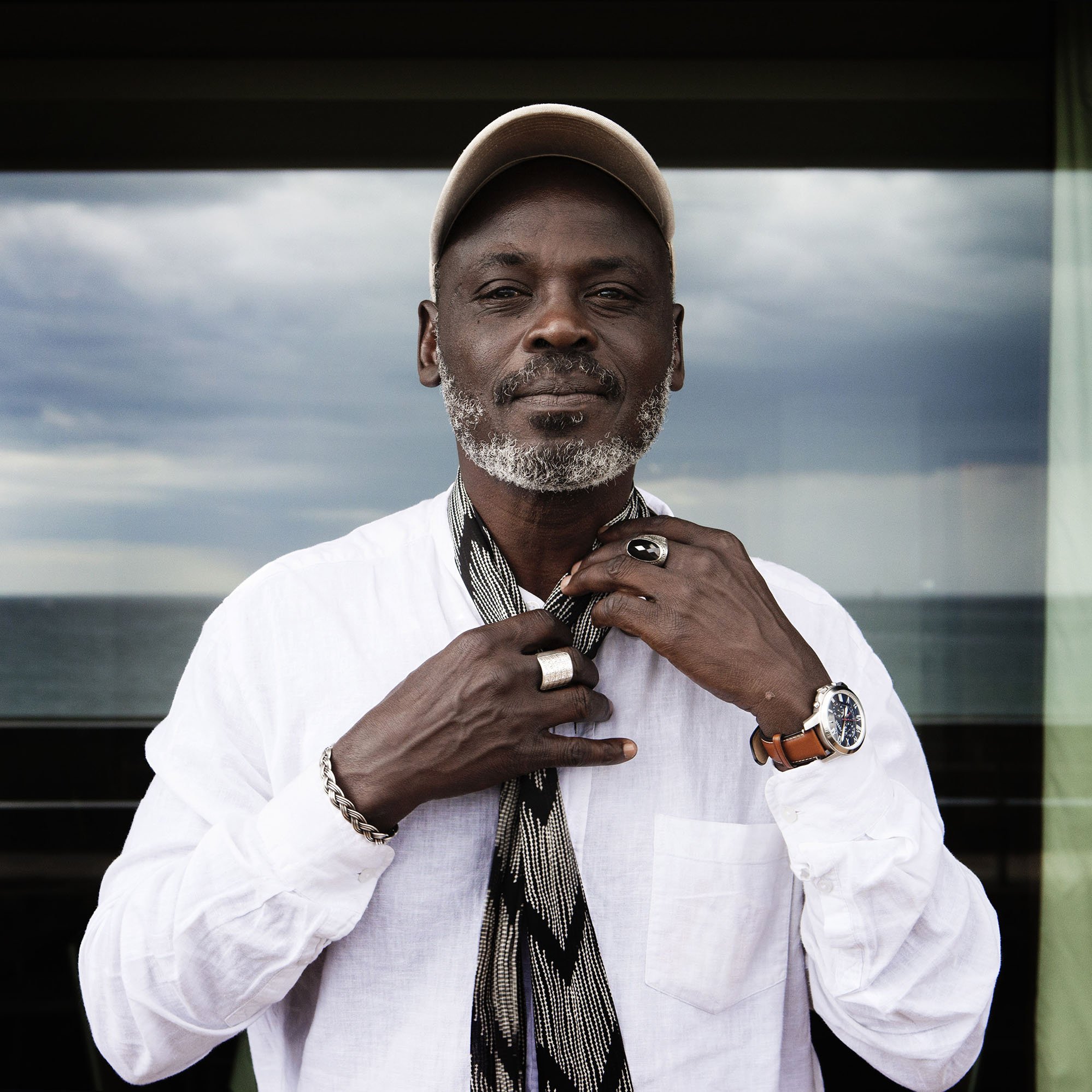 تظهر الصورة رجلًا يرتدي قميصًا أبيض وسلسلة ويضع قبعة على رأسه. يبدو أنه يثبت وشاحًا حول رقبته، ويقف أمام منظر طبيعي يضم البحر في الخلفية. السماء تظهر غائمة وكأنها تحمل بعض الغيوم. ملامح وجهه تعكس الثقة والهدوء.