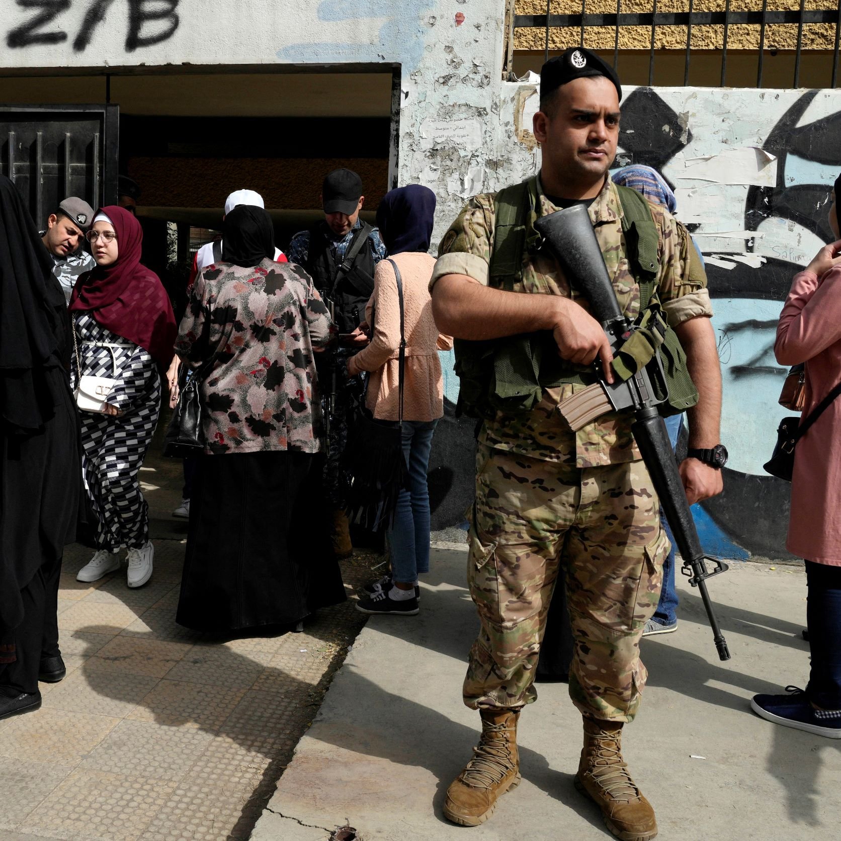 L'image montre un soldat armé en tenue militaire, se tenant debout et vigilant à l'extérieur d'un bâtiment. Au fond, plusieurs femmes sont rassemblées, certaines portant des hijabs et des vêtements de différentes couleurs. Le cadre semble urbain, avec des murs décorés de graffitis. L'atmosphère semble chargée de tension, avec la présence militaire suggérant un contexte de sécurité ou de contrôle.
