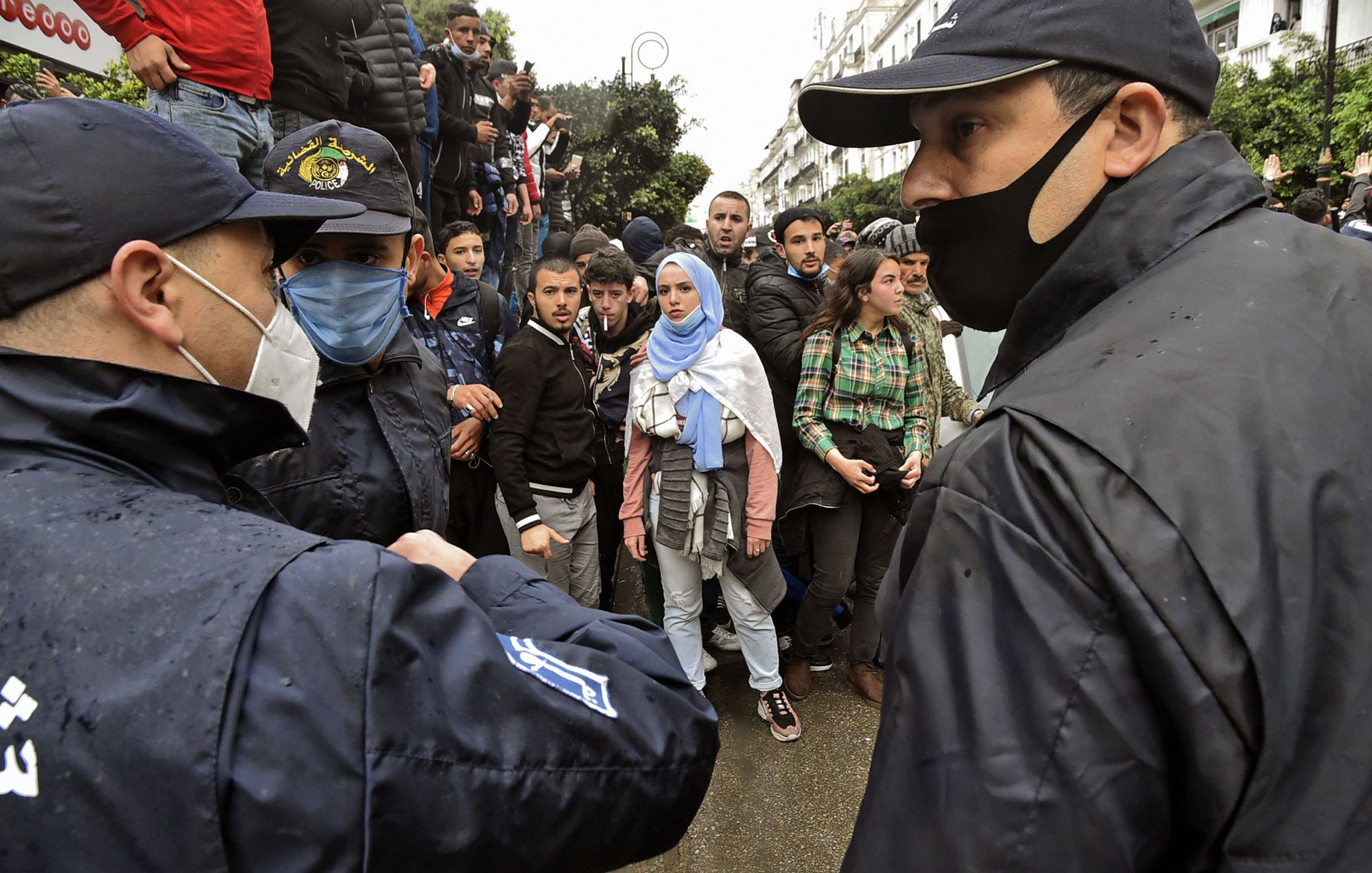 L'image montre une scène de tension entre des personnes en masse et des policiers. On peut apercevoir plusieurs individus, dont une femme portant un hijab et des vêtements clairs, qui semblent interagir avec les forces de l'ordre. Les policiers, en uniforme et portant des masques, sont en position d'encadrer la situation. L'arrière-plan montre une foule, indiquant un rassemblement ou une manifestation, et le temps semble pluvieux, ce qui ajoute à l'atmosphère de la scène.