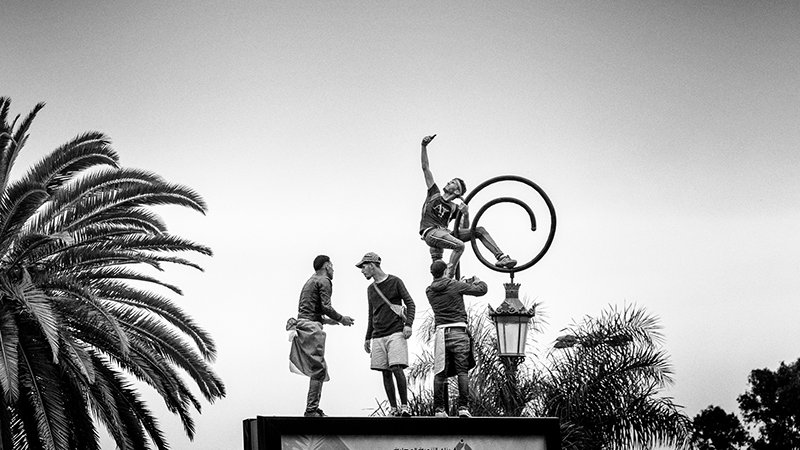 L'image montre un groupe de personnes en action, probablement en train de réaliser une performance acrobatique ou un numéro de cirque. L'une des personnes est perchée au sommet d'un cercle, tandis que les autres l'entourent, soutenant ou aidant dans la mise en scène. Le fond est dominé par un ciel clair et des palmiers, ce qui donne une ambiance estivale à la scène. L'ensemble est photographié en noir et blanc, ajoutant une touche artistique et dramatique.