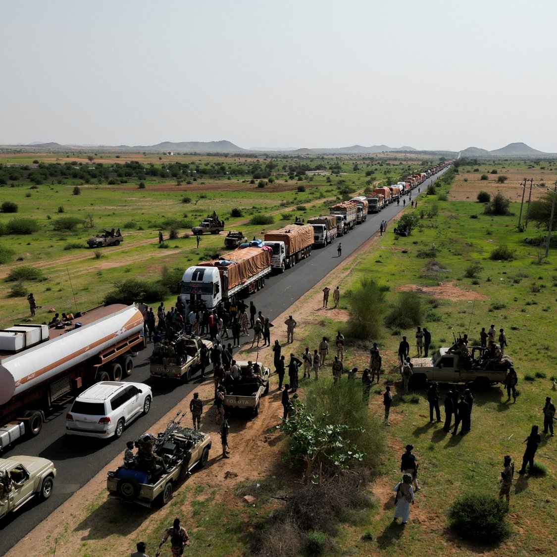 Convoi de camions sur une route, entouré de militaires et de verdure désertique.