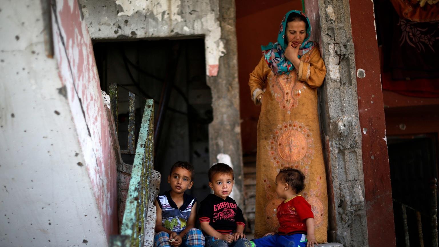 L'image montre une femme et plusieurs jeunes enfants assis sur des marches en béton. La femme est vêtue d'une robe longue, tandis que les enfants, assis près d'elle, portent des vêtements colorés. Le décor environnant semble ancien et en mauvais état, avec des murs écaillés et des escaliers. L'ambiance générale évoque une atmosphère de vie quotidienne dans un environnement modeste.