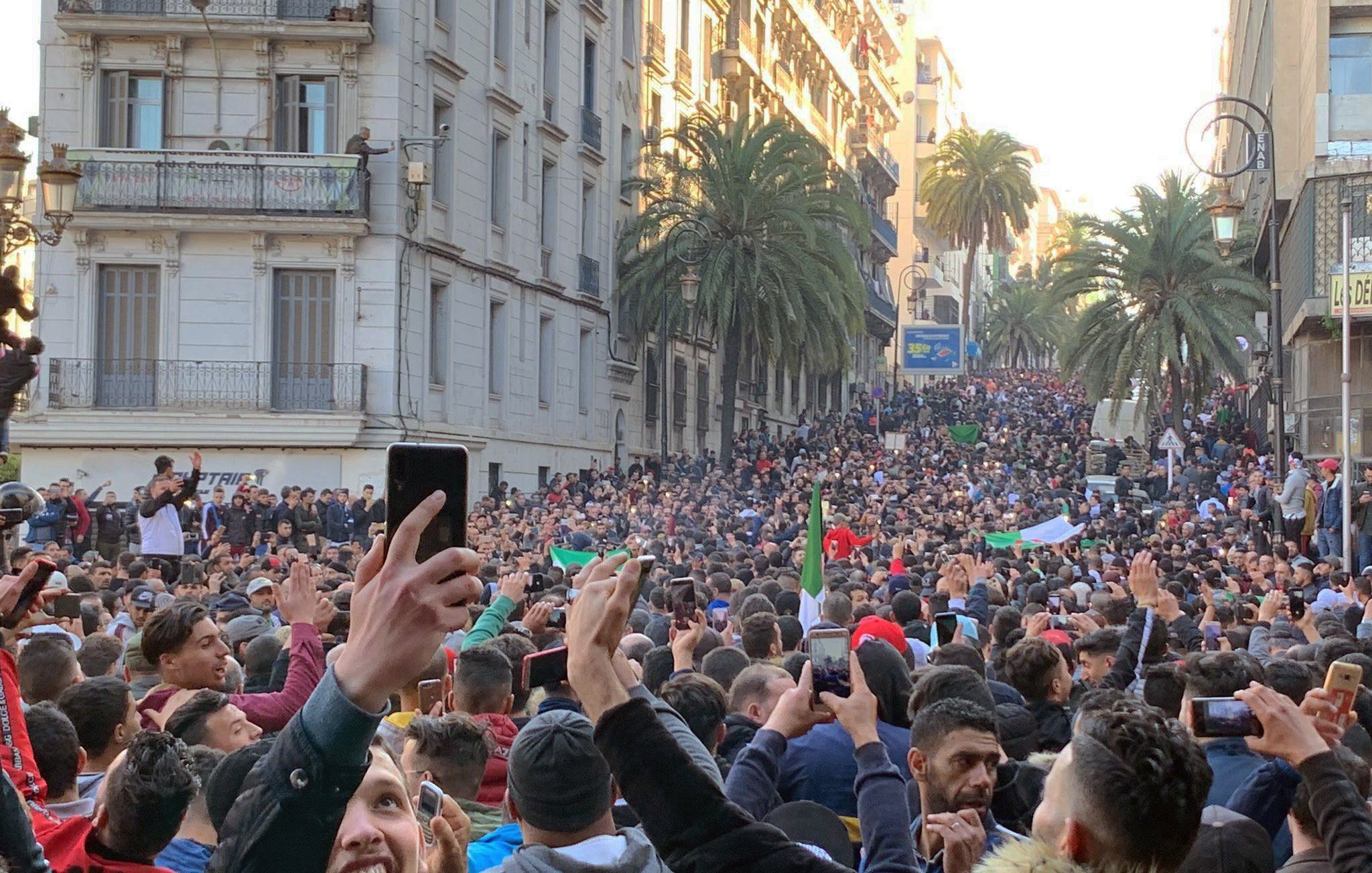 L'image montre une grande foule de personnes rassemblées dans une rue. Les participants semblent manifester, brandissant des drapeaux et levant des mains, certains tenant des téléphones pour filmer. On peut voir des bâtiments en arrière-plan, ainsi que des palmiers qui ajoutent une touche estivale à la scène. L'atmosphère semble dynamique et engagée, illustrant un moment de solidarité ou de protestation.