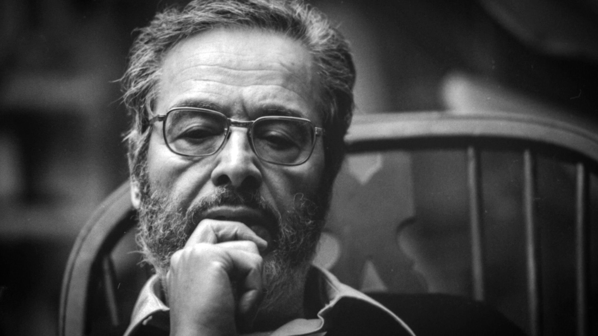 The image features a man with a thoughtful expression, sitting in a wooden chair. He has a beard and glasses, and he appears to be deep in contemplation. The lighting is soft, creating a moody atmosphere, and the background is blurred, focusing attention on his contemplative demeanor. The photograph has a classic black-and-white aesthetic, emphasizing the seriousness of the moment.