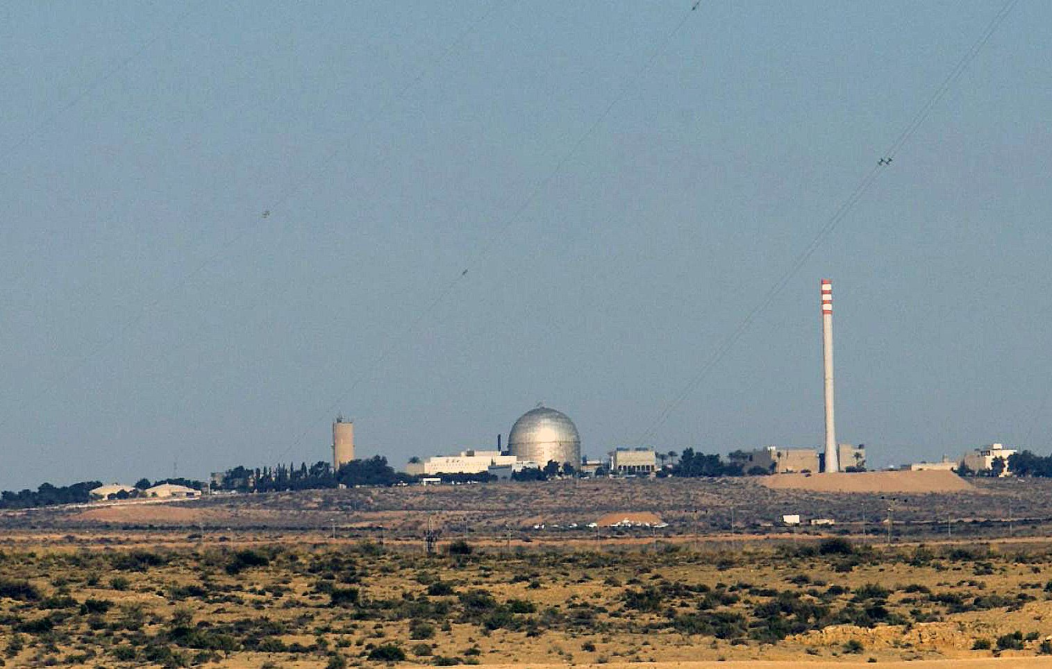 The image depicts a desert landscape with a prominent facility in the background. This facility features a large, domed structure, likely indicative of a scientific or research establishment, alongside tall smokestacks or cooling towers. The setting appears arid, with sparse vegetation, and the horizon stretches into the distance, emphasizing the isolation of the facility. The overall atmosphere conveys a sense of industrial or technological presence amidst a natural environment.