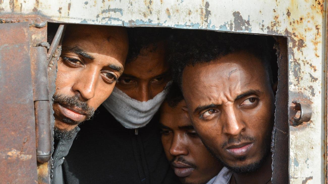 تظهر في الصورة مجموعة من الأشخاص ينظرون من خلال فتحة صغيرة في باب أو جدار صدئ. تعكس تعابير وجوههم مشاعر القلق والترقب، كما يبدو أنهم مقيدون أو محاطون بظروف صعبة. الخلفية تظهر علامات التآكل والصدأ، مما يضيف إلى الأجواء العامة للصورة.