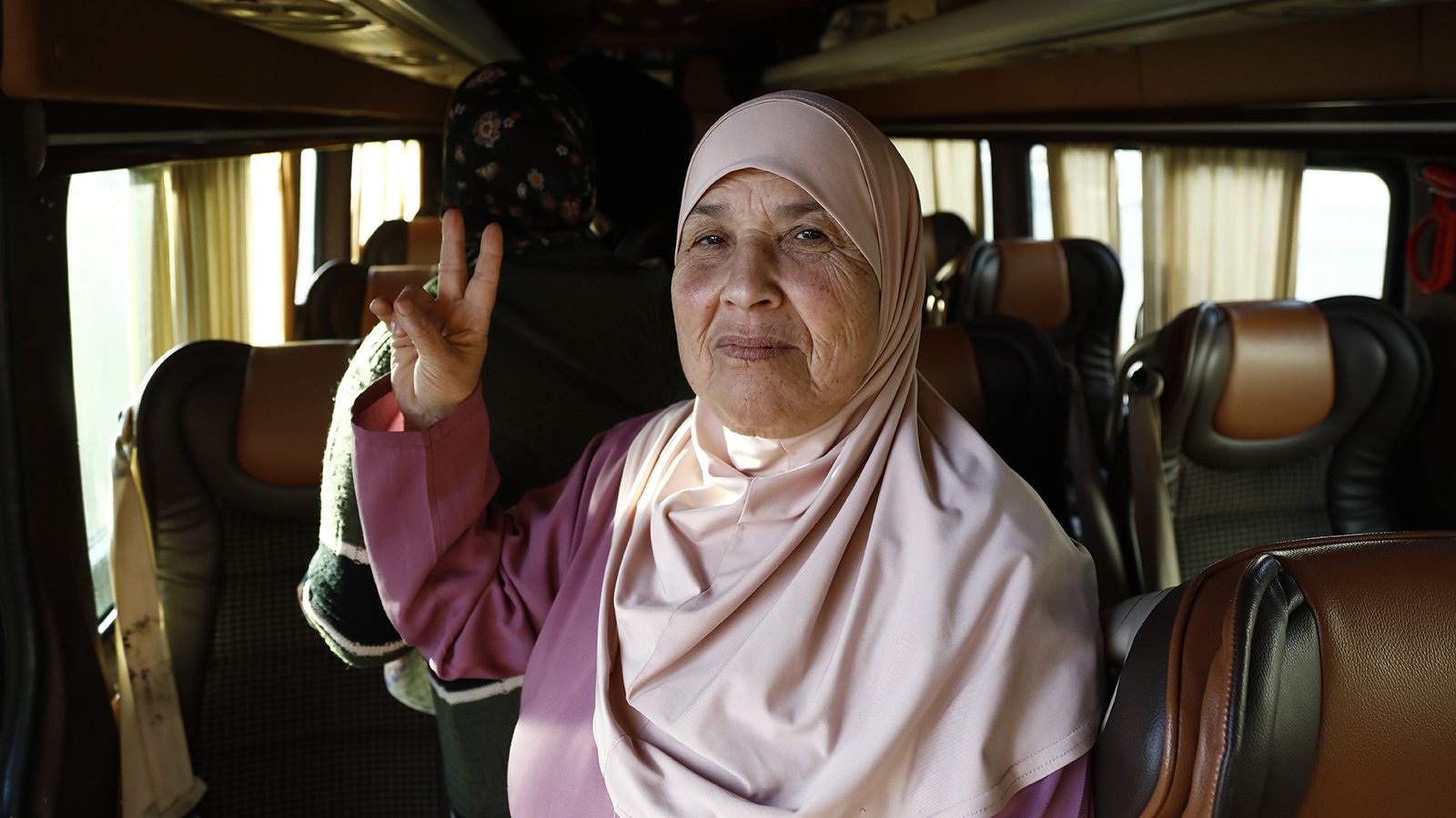 الصورة تظهر امرأة مسنّة ترتدي حجابًا بلون وردي. تبدو مبتسمة وتلوح بإشارة النصر بإصبعين. يبدو أنها داخل حافلة، مع مقاعد مريحة ورؤية لنساء أخريات في الخلفية. تعكس الصورة جوًّا من الألفة والفرح.