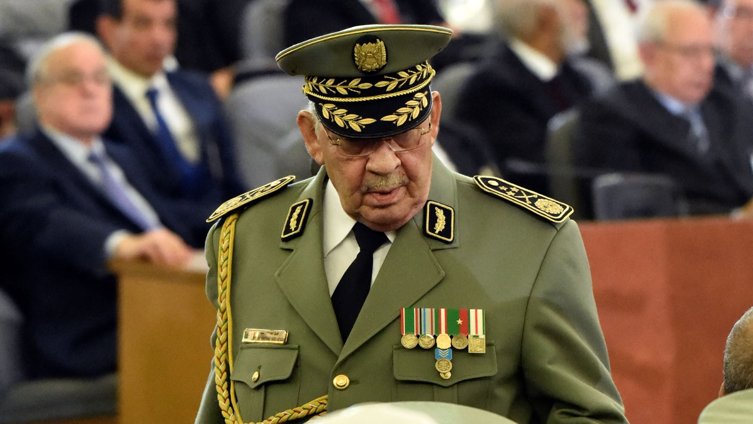 L'image montre un groupe de personnes réunies dans une grande salle, avec des sièges disposés en rangées. Au premier plan, un homme en uniforme militaire avec un chapeau prononcé, orné de médailles, se déplace parmi la foule. Dans l'arrière-plan, on aperçoit d'autres hommes et femmes, certains en costume, d'autres portant des turbans, tous attentifs à l'événement. L'atmosphère semble formelle, suggérant une occasion importante ou une cérémonie.