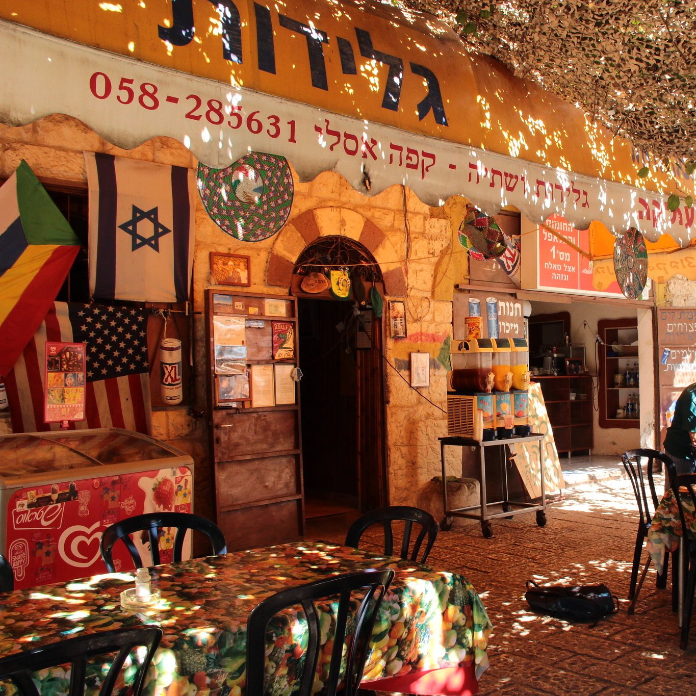الصورة تظهر مقهى أو مطعمًا يحتوي على عدد من الطاولات والكراسي، ويبدو أن المكان مفتوح وتغطيه مظلة من الأوراق أو النباتات. الأجواء تبدو مريحة ومرحب بها، حيث يجلس عدد من الزبائن في أماكنهم. هناك لافتات باللغتين العبرية والإنجليزية، كما تظهر أعلام متنوعة، مما يدل على تنوع الزوار. يمتد المكان في منطقة جمجومية، مع ضوء الشمس يتسلل من خلال الأوراق.