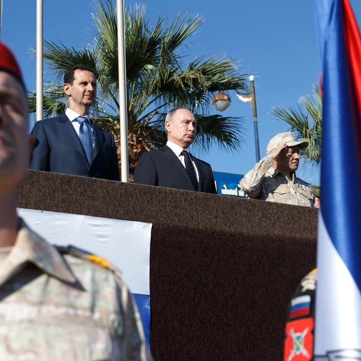 The image depicts a military ceremony or event, featuring several individuals of importance. In the foreground, soldiers in military uniforms with red berets are standing at attention. In the background, a group of officials is present, including two prominent figures positioned together: one in a suit and tie and the other in military attire. There's a large flag in the foreground, possibly representing a nation involved in the event. The setting appears outdoors, with palm trees visible, suggesting a warm climate. The overall atmosphere seems formal and significant, likely indicative of a diplomatic or military alliance.