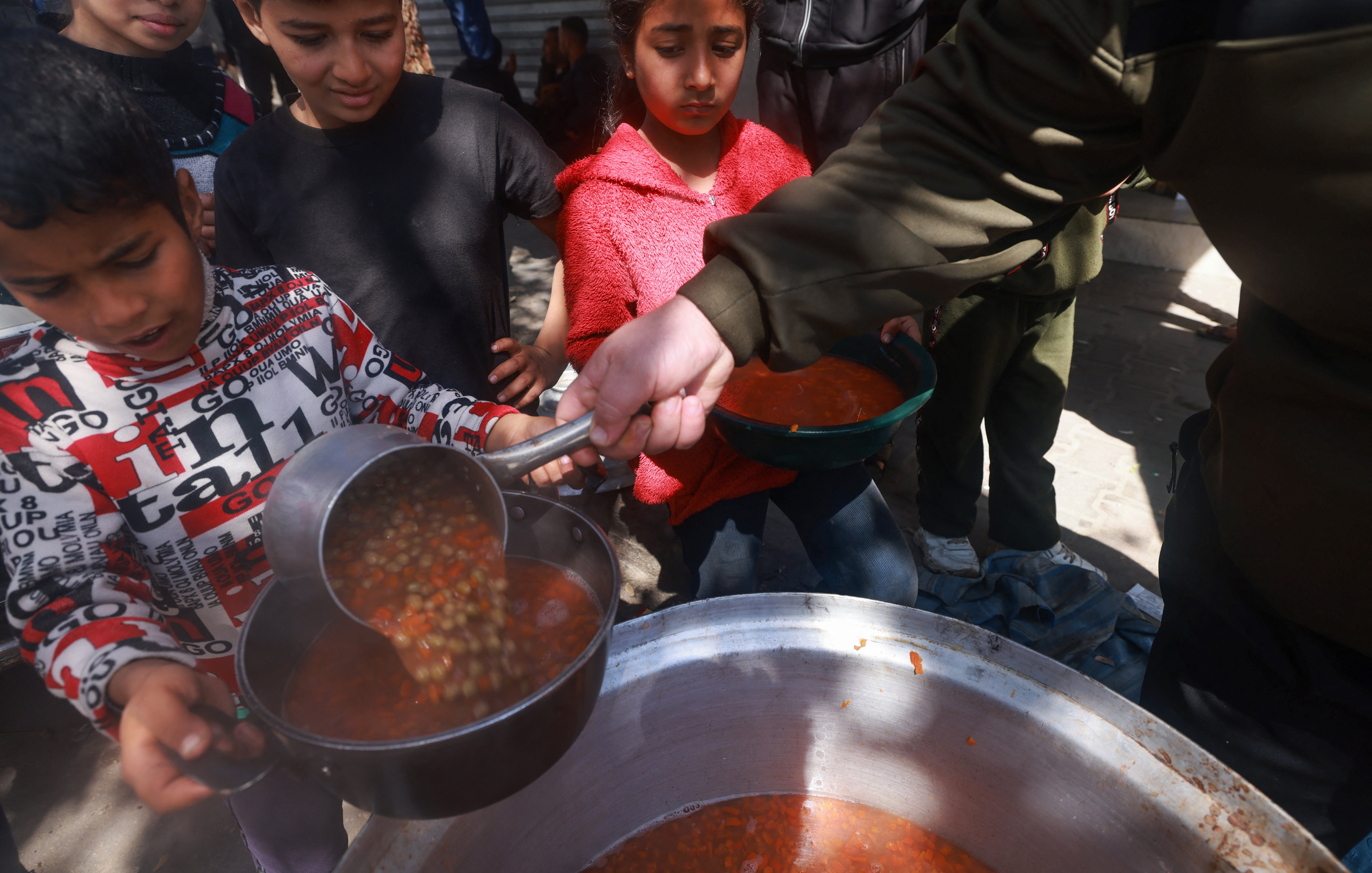 L'image montre des enfants qui se rassemblent autour d'une grande marmite. Un enfant, tenant une louche, verse une soupe dans un bol. D'autres enfants, attentifs, attendent leur tour pour recevoir leur repas. L'atmosphère semble conviviale, et l'environnement est extérieur, probablement dans une aire où des repas sont servis. Les enfants portent des vêtements variés, et certains semblent impatients et enthousiastes.