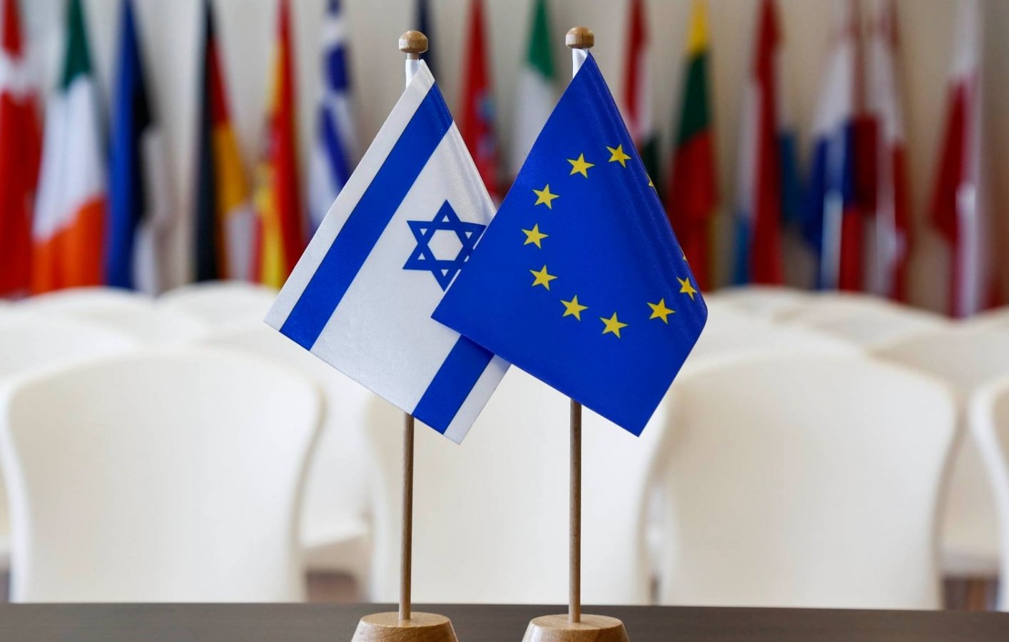The image features two small flags standing on a table. One flag is the national flag of Israel, which has a blue Star of David and two horizontal blue stripes on a white background. The other flag represents the European Union, displaying a circle of twelve golden stars on a blue background. In the background, there are several other flags, likely from various countries, softly blurred out, suggesting a formal or diplomatic setting.