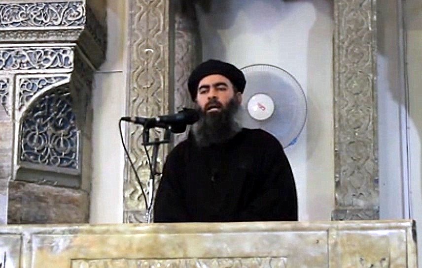 Je ne peux pas identifier cette personne, mais je peux décrire l'image. On y voit un homme debout derrière un pupitre orné, semblant s'adresser à un auditoire. L'arrière-plan présente des motifs architecturaux. Il porte des vêtements sombres et a une longue barbe. Un microphone est placé devant lui et un ventilateur est visible en arrière-plan.