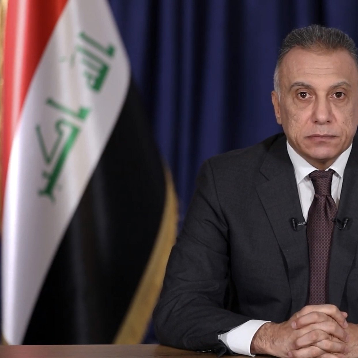 در این تصویر، مردی در حال نشسته با ظاهری رسمی دیده می‌شود. او لباس تیره‌ای به تن دارد و کراواتی به رنگ تیره نیز بر گردن دارد. چهره‌اش جدی و متمرکز است و دستانش به صورت مرتب روی میز مقابلش قرار گرفته‌اند. در پس‌زمینه، پرده‌ای با رنگ تیره وجود دارد و همچنین پرچم عراق در سمت چپ تصویر نمایان است. این تصویر به نظر می‌رسد که به یک سخنرانی یا مصاحبه رسمی مربوط باشد.
