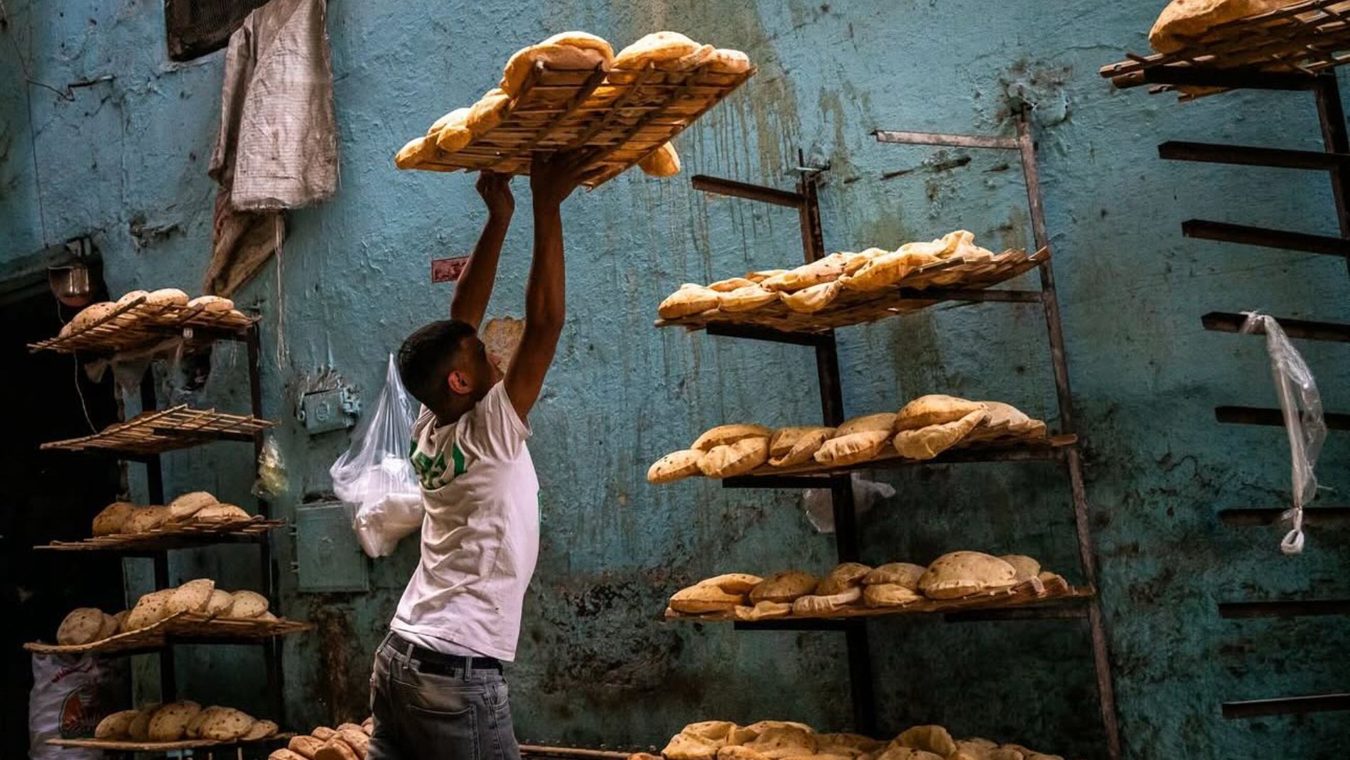 Un boulanger soulève un plateau de pains dans une boulangerie sur un mur bleu.