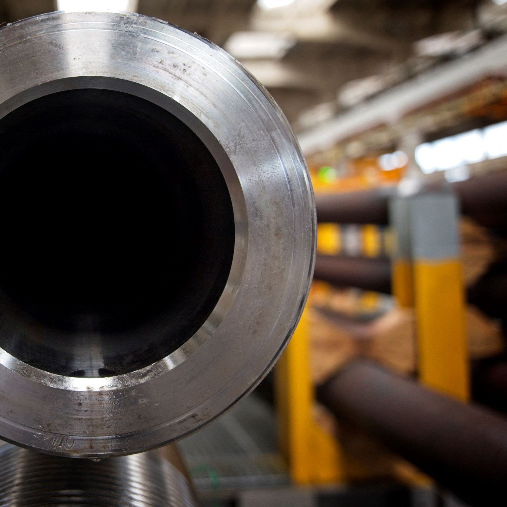 La imagen muestra el extremo de un tubo de metal, donde se puede ver su interior oscuro y liso. El tubo tiene un acabado brillante en su parte exterior, y el entorno sugiere un ambiente industrial, con otros tubos y estructuras a lo lejos, pero enfocados en primero plano. La iluminación es suave y resalta los detalles del material.