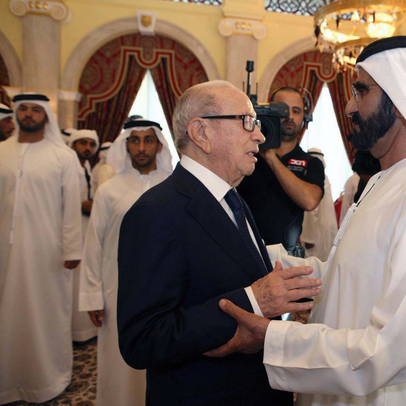 الصورة تُظهر شخصين يتبادلان الحديث في مكان فخم مزين بألوان دافئة وزخارف تقليدية. يبدو أن أحدهما يرتدي بدلة رسمية بينما الآخر يرتدي الزي الإماراتي التقليدي. يُحيط بهما عدد من الأشخاص في الخلفية، والجو العام يوحي بأجواء من الحفاوة والترحيب.