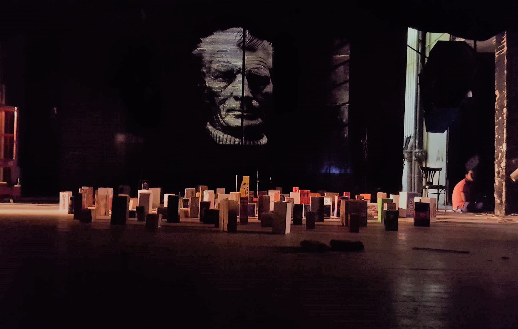 L'image montre une scène intérieure avec un grand mur sombre sur lequel se trouve un portrait en noir et blanc d'une personne âgée. Au premier plan, il y a un sol rempli de petites structures ressemblant à des livres ou des modules disposés de manière organisée. À l'arrière-plan, on peut apercevoir une personne assise, probablement en train de travailler ou de se préparer. L'éclairage est faible, créant une atmosphère mystérieuse.