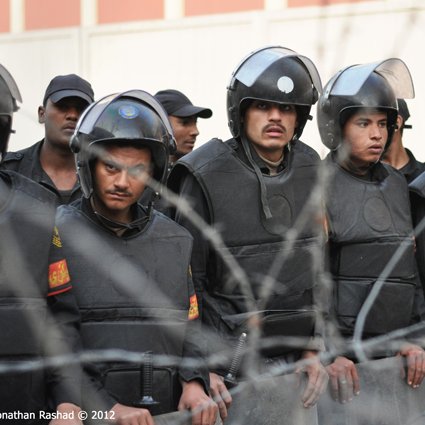 L'image montre un groupe de policiers en tenue anti-émeute, portant des casques et des gilets pare-balles. Ils semblent être en formation et regardent attentivement dans une direction, manifestant une posture de vigilance. En arrière-plan, on peut voir des barbelés, ce qui indique un contexte de sécurité renforcée ou de tensions. Les soldats portent des uniformes sombres et sont alignés, ajoutant à l'impression de discipline et de préparation.