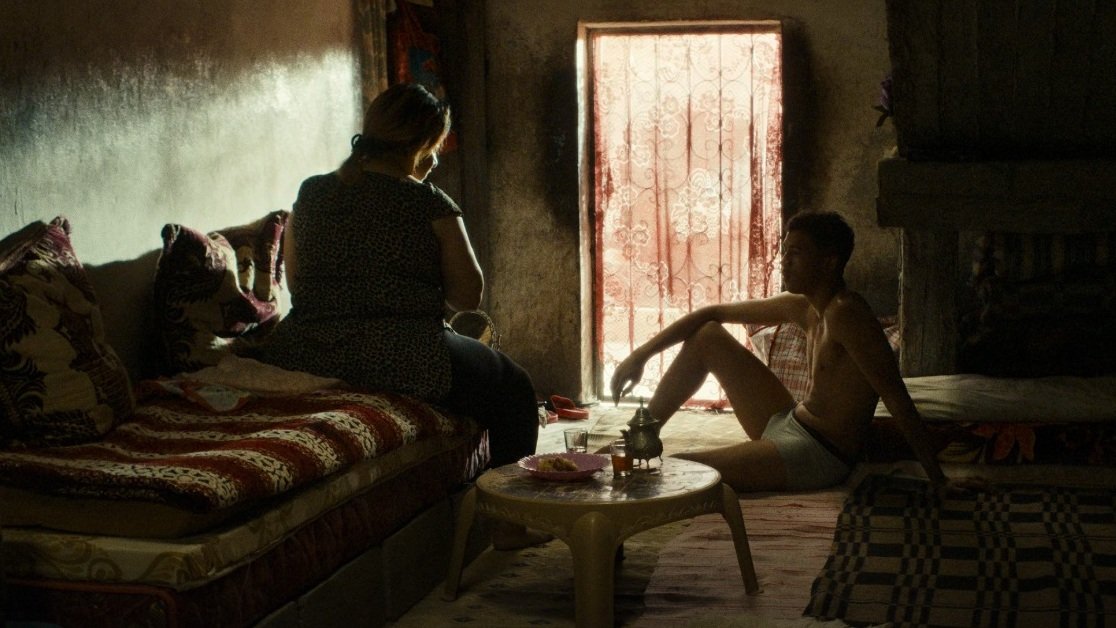 L'image montre une scène intérieure, probablement dans un espace de vie modeste. On y voit deux personnes, assises dans une pièce faiblement éclairée par la lumière qui entre à travers une fenêtre avec des rideaux. Les murs sont de couleur sombre et le mobilier semble simple. L'une des personnes est assise sur un canapé, tandis que l'autre est assise par terre, semblant contempler la pièce ou se relaxer. L'atmosphère dégage une sensation d'intimité et de calme. Des objets sont présents, suggérant une vie quotidienne.