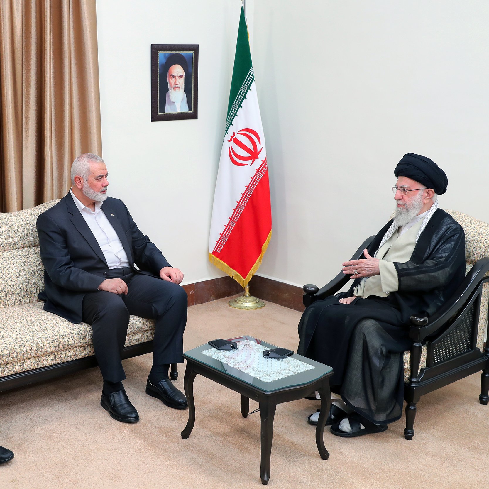 L'immagine mostra un incontro tra tre uomini. Due di loro sono seduti su un divano, mentre il terzo, che indossa un abbigliamento tradizionale, è seduto di fronte a loro. Sul muro si trova un ritratto e, accanto, è presente una bandiera iraniana. L'atmosfera sembra formale e il gruppo appare impegnato in una discussione.