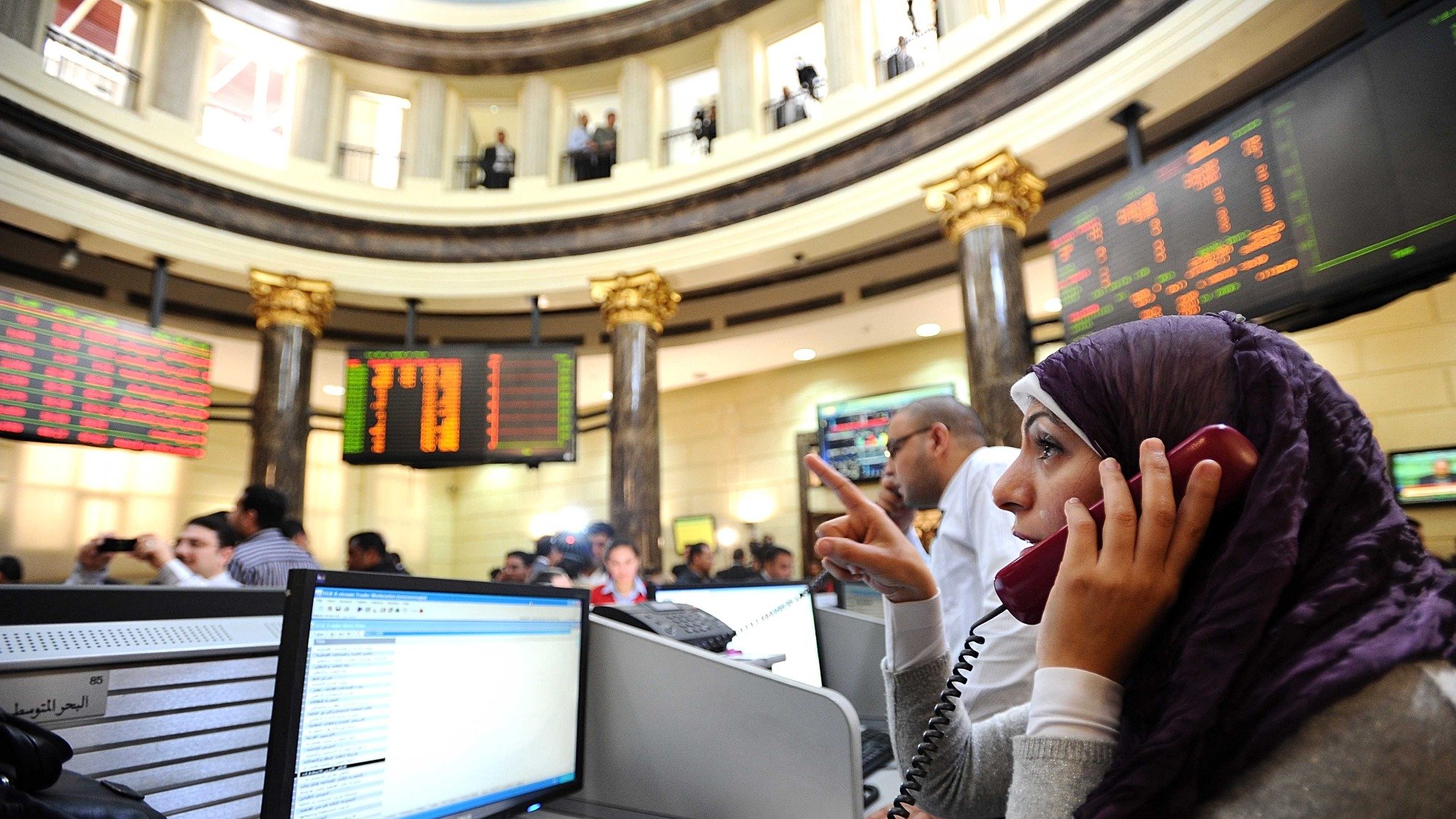 تظهر الصورة مشهداً من داخل سوق الأسهم، حيث يوجد عدد من الأشخاص يتعاملون مع شاشات تعرض الأسعار والبيانات المالية. في المقدمة، توجد امرأة ترتدي حجاباً، وهي تتحدث عبر الهاتف. يبدو أنها مشغولة بالعمل، حيث تشير بإحدى يديها. الخلفية تحتوي على عدة أشخاص آخرين وشاشات تعرض معلومات السوق. الأجواء توحي بالجدية والنشاط المالي.