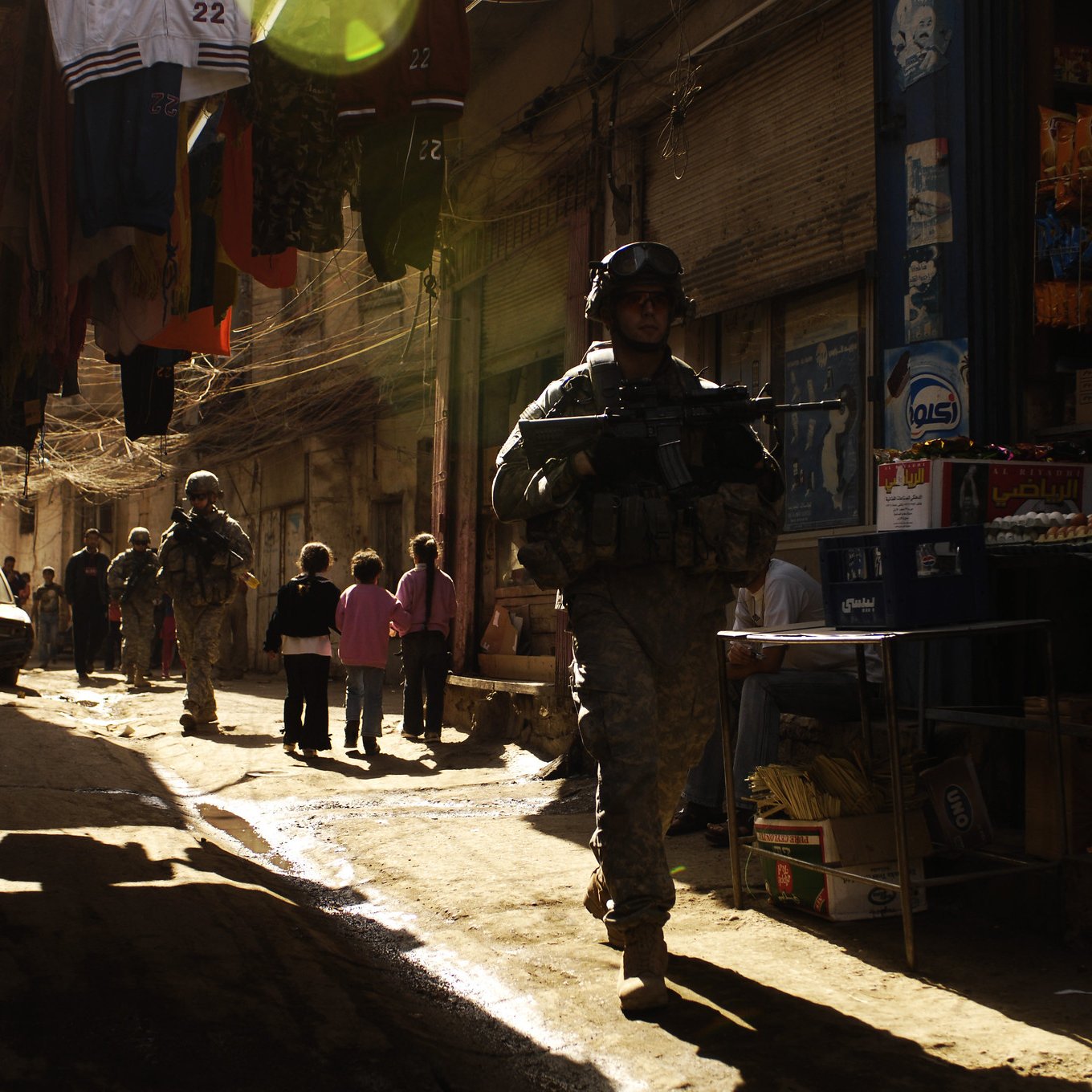 La imagen muestra a soldados patrullando en un callejón estrecho y iluminado por la luz del sol. A los lados, hay ropa colgada y tiendas de barrio que parecen ofrecer diversos productos. Un grupo de niños está presente en la escena, observando a los soldados. La atmósfera es tensa, dado el contexto militar, pero la luz crea un efecto visual interesante en el entorno.