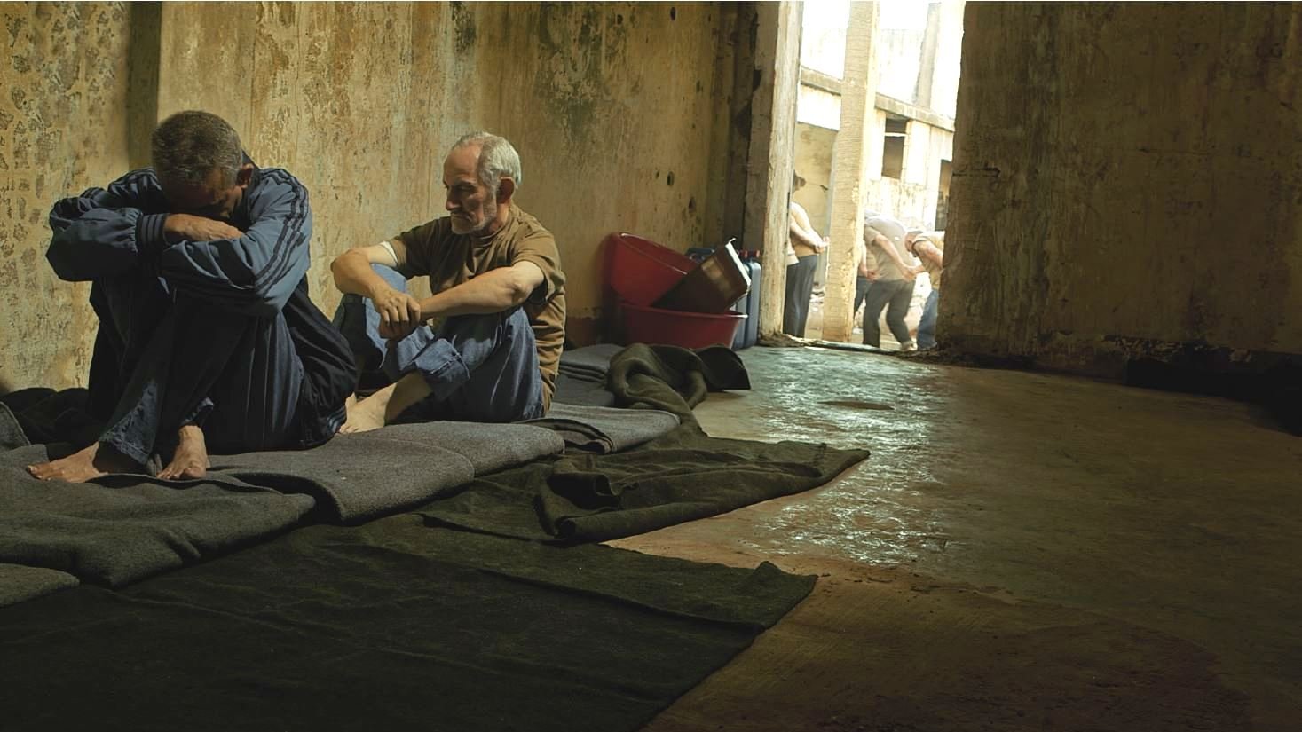 L'image montre un espace intérieur sombre et désolé, probablement une cellule ou un lieu de détention. À l'avant, deux hommes sont assis sur des couvertures posées sur le sol. L'un semble pensif et acculé, tandis que l'autre est plus détendu et semble essayer de communiquer ou de réconforter. En arrière-plan, d'autres personnes sont visibles, marchant en file. L'éclairage est faible, ce qui contribue à une atmosphère de tristesse et de confinement. Les murs semblent usés et abandonnés, renforçant l'impression de désespoir.