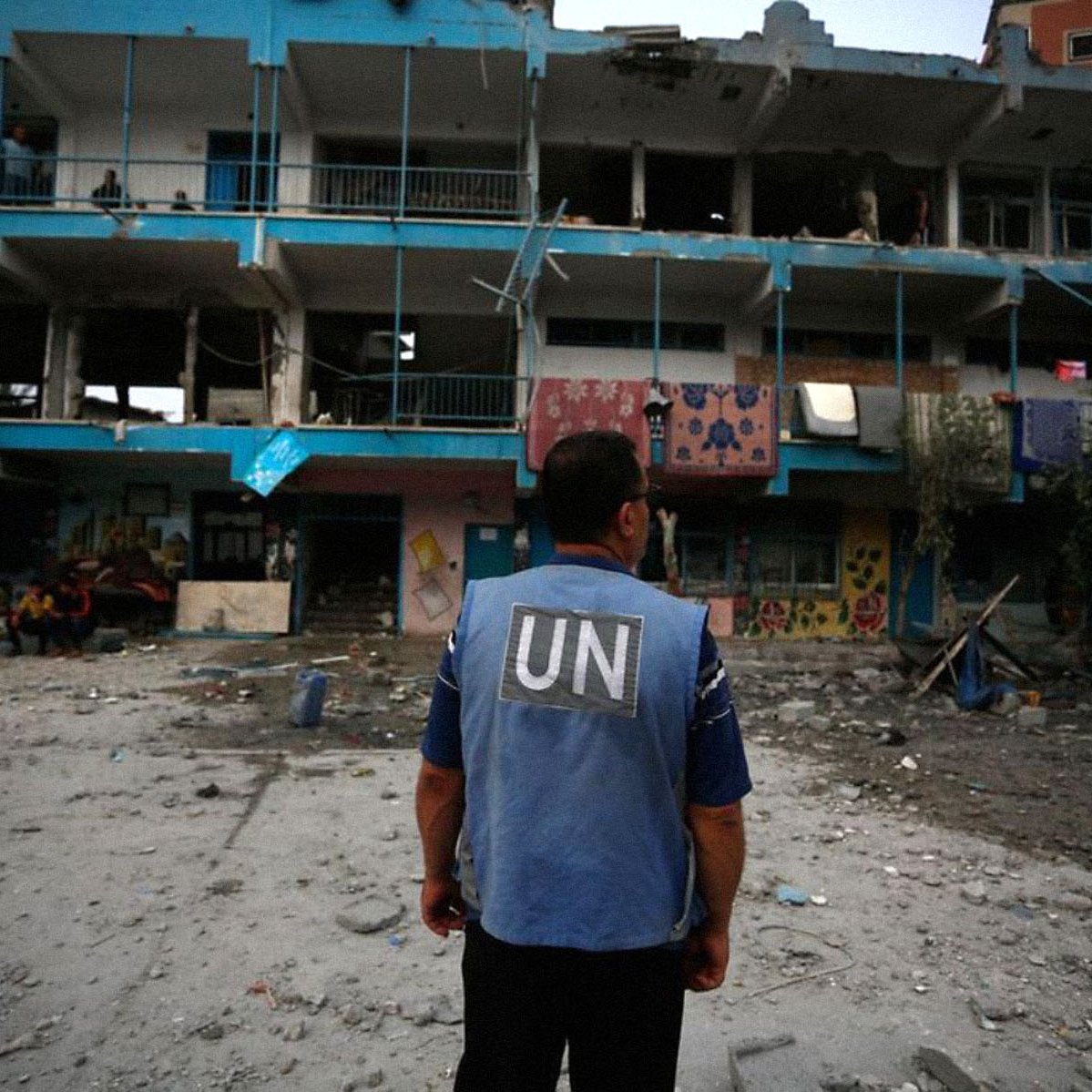 L'image montre un homme portant un gilet de l'ONU, observant un bâtiment endommagé. Le bâtiment a des murs peints et des balcons, mais il est en ruine, avec des fenêtres brisées et des débris éparpillés autour. L'atmosphère est sombre, suggérant une situation de conflit ou de crise, avec d'autres personnes visibles en arrière-plan dans un environnement chaotique.