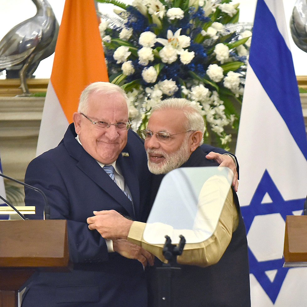 L'image montre deux hommes en train de se serrer dans les bras, exprimant une atmosphère chaleureuse et amicale. L'un des hommes porte un costume sombre avec une cravate, tandis que l'autre est vêtu d'une tenue traditionnelle indienne. En arrière-plan, on peut voir des drapeaux de l'Inde et d'Israël ainsi que des fleurs décoratives, ce qui indique un contexte officiel ou diplomatique.