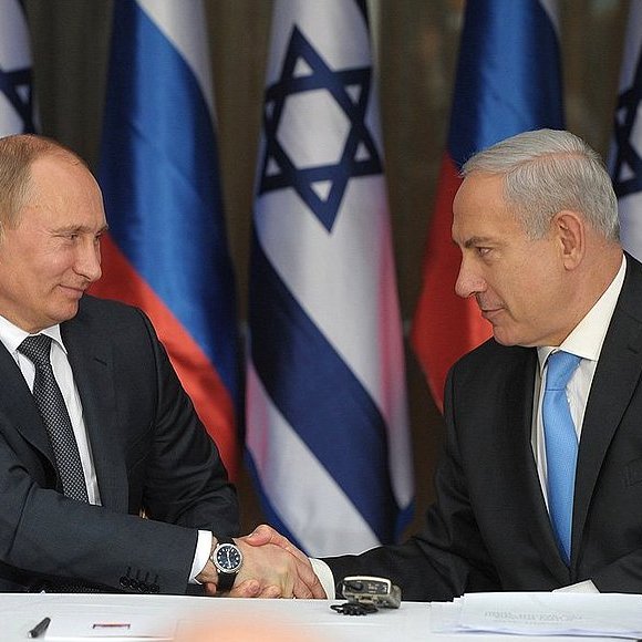 L'image montre deux hommes en costume, se serrant la main, dans un cadre formel. À leur arrière-plan, on peut voir des drapeaux de la Russie et d'Israël. L'un des hommes, à gauche, semble être en train de porter un écouteur, tandis que l'autre, à droite, regarde attentivement. L'ambiance suggère un moment de coopération ou d'accord.