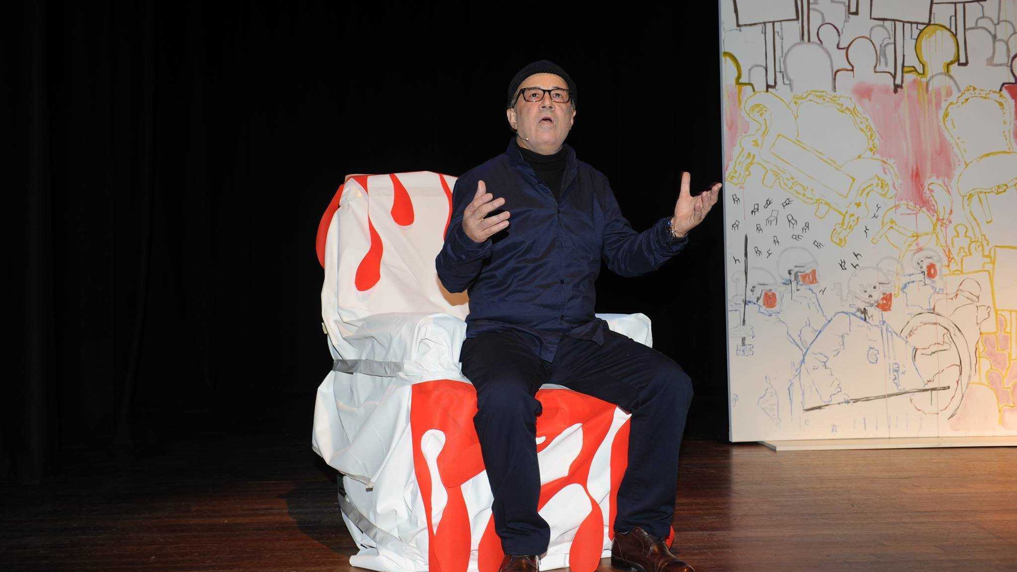 The image features a man sitting on a uniquely designed chair that has a colorful, artistic pattern, predominantly featuring red and white. He is wearing a dark outfit, including a jacket and glasses, and appears to be expressing himself with his hands raised, as if making a point during a performance or presentation. The background includes a large, stylized backdrop with a simple, colorful design. The setting seems to be a stage, suggesting a theatrical or artistic context.
