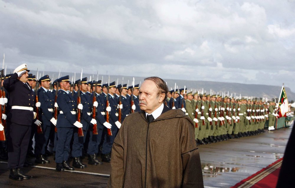 L'image montre un homme en manteau se tenant devant un alignement de soldats en uniforme. Les soldats, majestueux et bien rangés, saluent. L'arrière-plan est nuageux, créant une atmosphère cérémonielle. Le sol semble humide, indiquant peut-être qu'il a plu récemment. L'homme au premier plan a une expression pensive, ajoutant un ton sérieux à la scène.