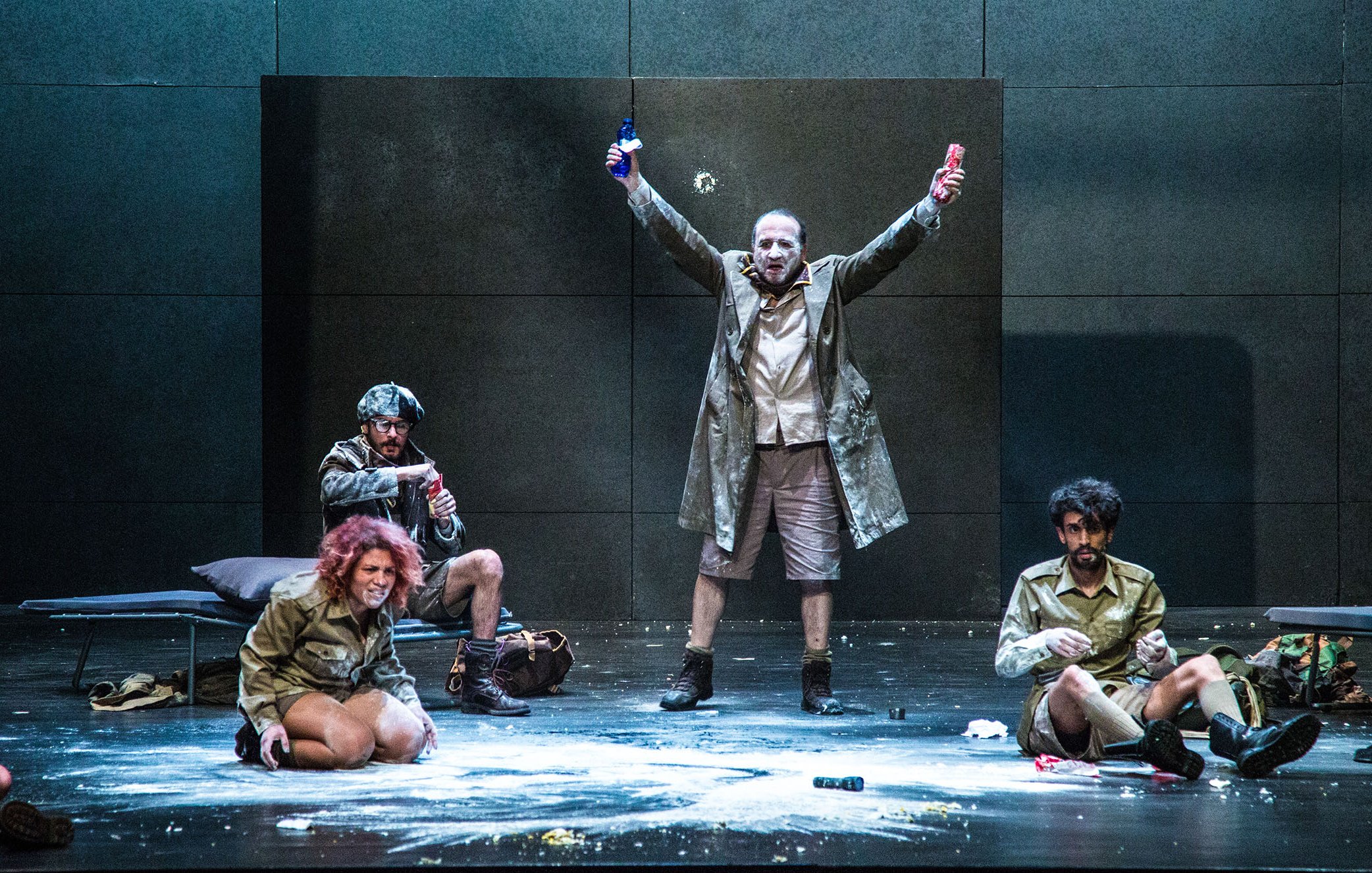 L'image montre une scène de théâtre sur une scène dépouillée. Au centre, un homme en manteau se tient avec les bras levés, tenant une bouteille dans chaque main, ce qui suggère une attitude triomphante ou exubérante. Autour de lui, plusieurs autres personnages sont assis ou accroupis, vêtus de costumes similaires. L'un d'eux semble concentré sur une activité au sol. La scène peut évoquer une ambiance dramatique ou absurde, avec des éléments de désordre comme de la poudre dispersée sur le sol. Les expressions des personnages varient, ajoutant à la dynamique de l'ensemble.