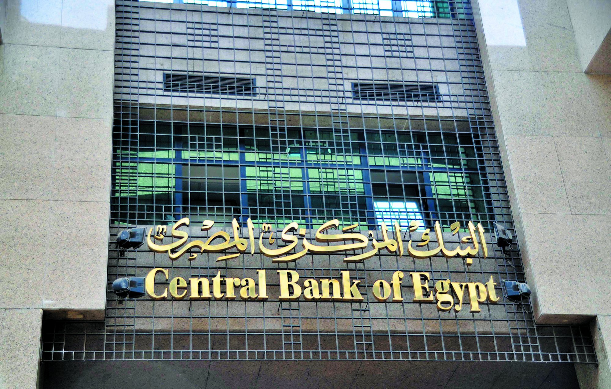 The image features the facade of a building that houses the Central Bank of Egypt. The building has a modern design with a prominent sign displaying "Central Bank of Egypt" in both English and Arabic. The Arabic text is situated above the English, and the overall appearance conveys a sense of official importance and stability. The building is constructed with stone materials, and its facade includes grid-like elements, suggesting a blend of contemporary and traditional architectural styles.