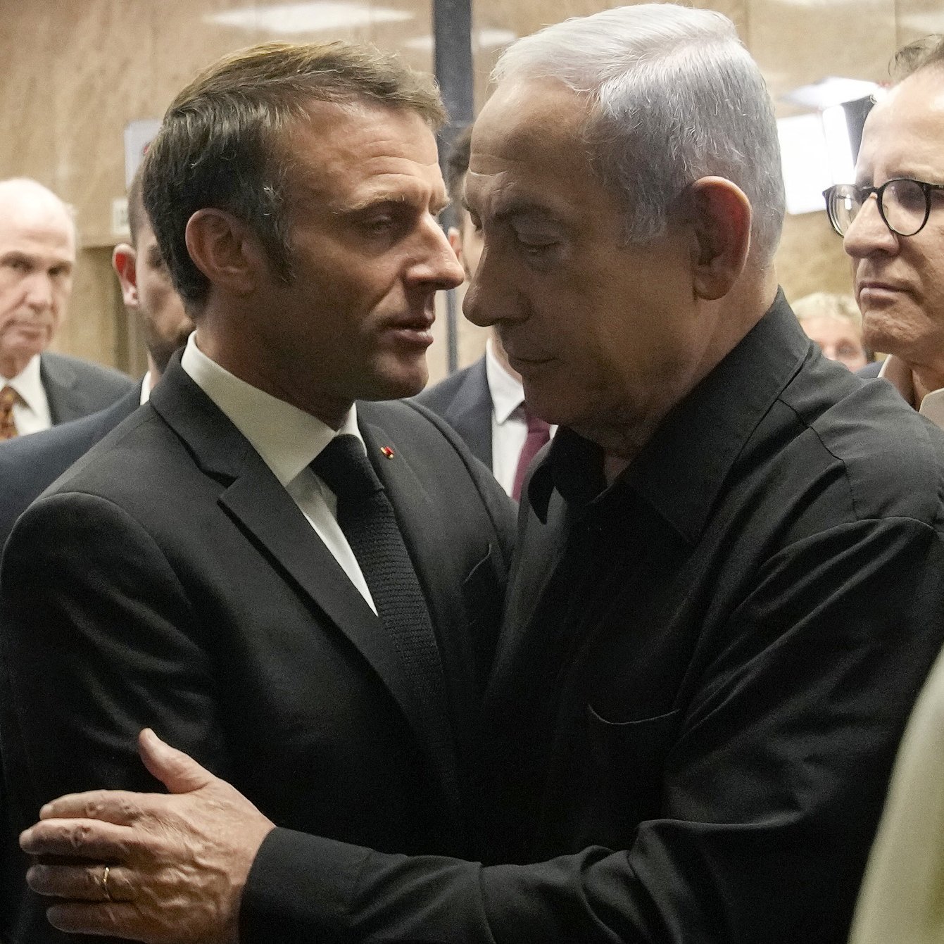 La imagen muestra a dos hombres en un entorno formal, aparentemente en un edificio gubernamental. Uno de ellos viste un traje oscuro y tiene una expresión seria mientras se acerca al otro, quien lleva una camisa negra. Ambos parecen en una conversación cercana, rodeados por varias personas que observan la interacción. La atmósfera sugiere un momento significativo, posiblemente de diálogo o entendimiento.