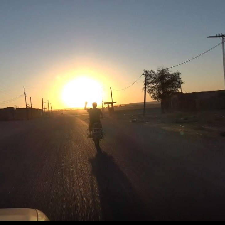 The image depicts a scenic view of a road extending into the distance during sunset. A person on a motorcycle is riding away from the camera, with their hand raised, possibly in a wave or gesture. The sun is setting on the horizon, casting a warm golden light across the landscape. On either side of the road, there are silhouettes of buildings and trees, and the scene conveys a sense of tranquility and adventure. Utility poles line the road, contributing to the rural setting.